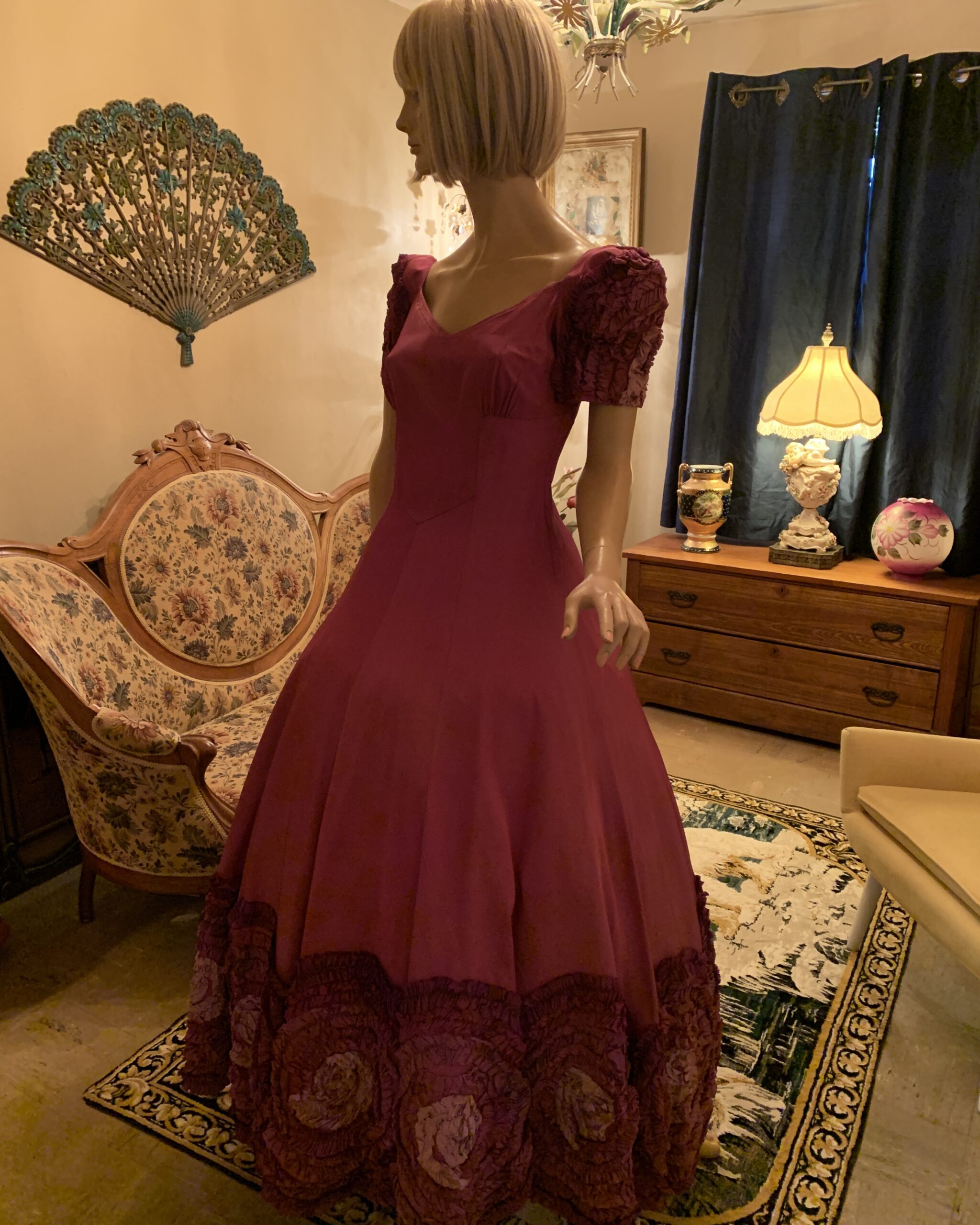 Magenta vintage evening gown displayed on mannequin with fitted bodice, sculpted waist, short textured sleeves, and dimensional rosette hem detail.