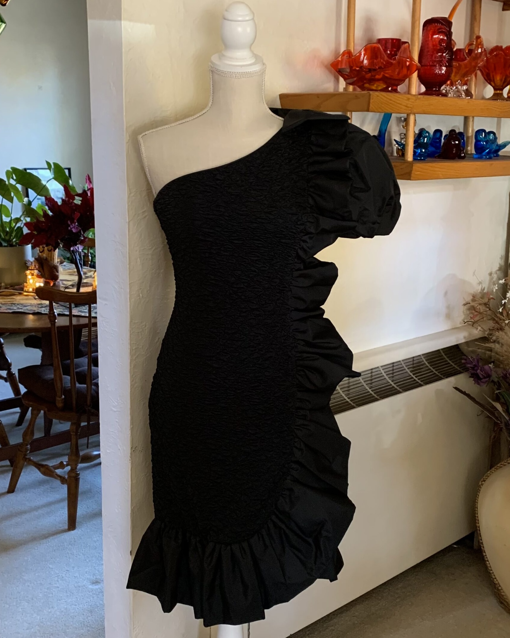 Black one-shoulder vintage cocktail dress with dramatic sculptural ruffles, textured ruched fabric, and rhinestone buckle detail displayed on a mannequin in a softly lit interior.