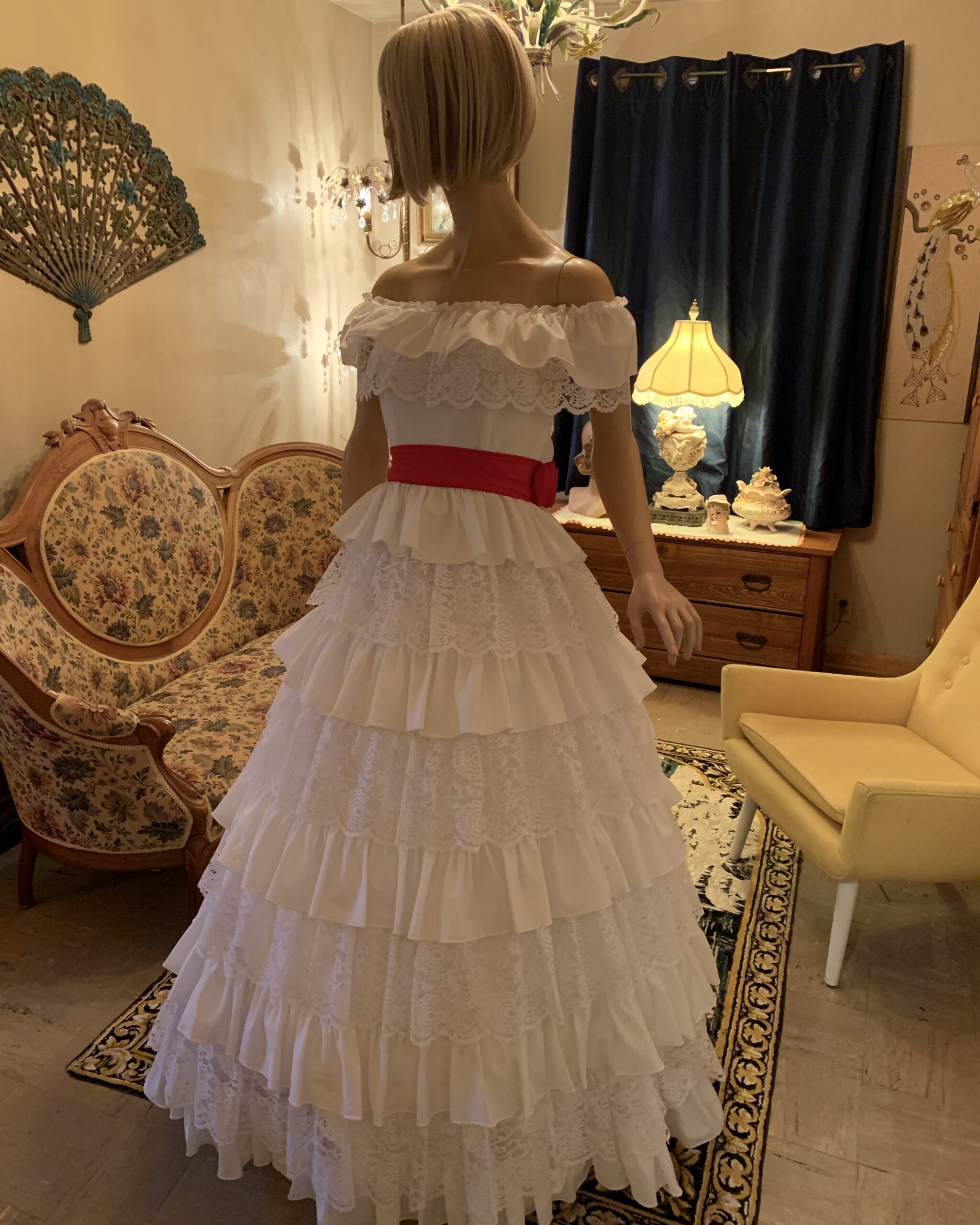 Three-quarter view of a vintage white lace gown with tiered ruffles, off-the-shoulder neckline, and fitted waist accented by a red sash.