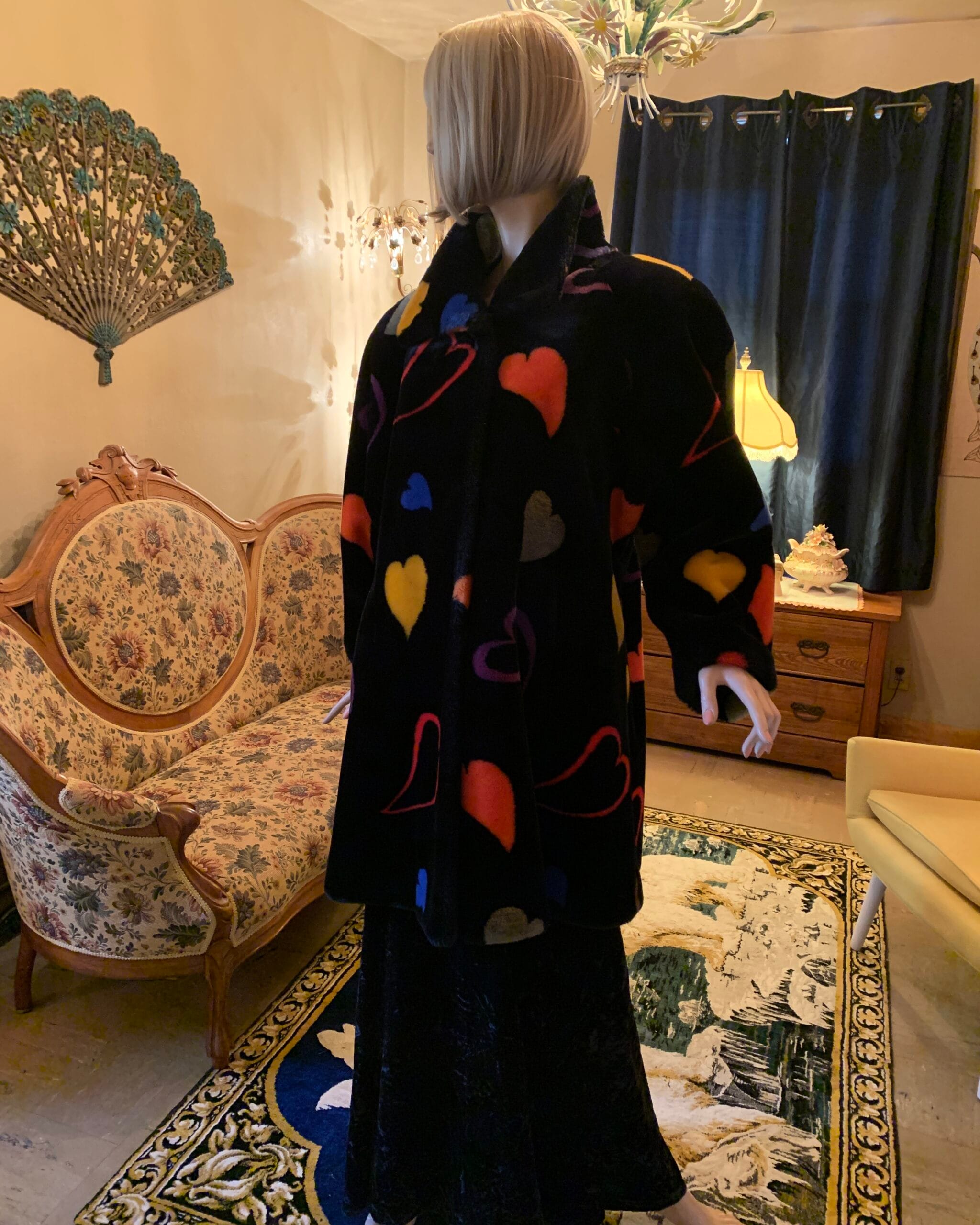 Side view of a vintage Donnybrook black plush coat with oversized multicolored hearts, displayed on a mannequin to highlight the drape, collar, and hidden front closure.