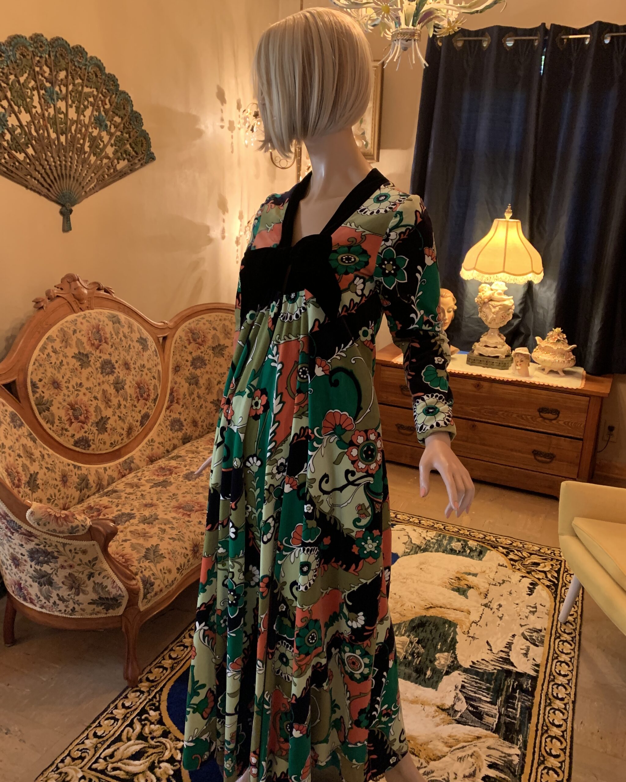 Vintage Bards Toronto maxi dress in a bold floral print of olive green, emerald, coral, cream, and black, featuring a dramatic black velvet bow at the empire waist and long sleeves, displayed in a vintage-inspired salon.