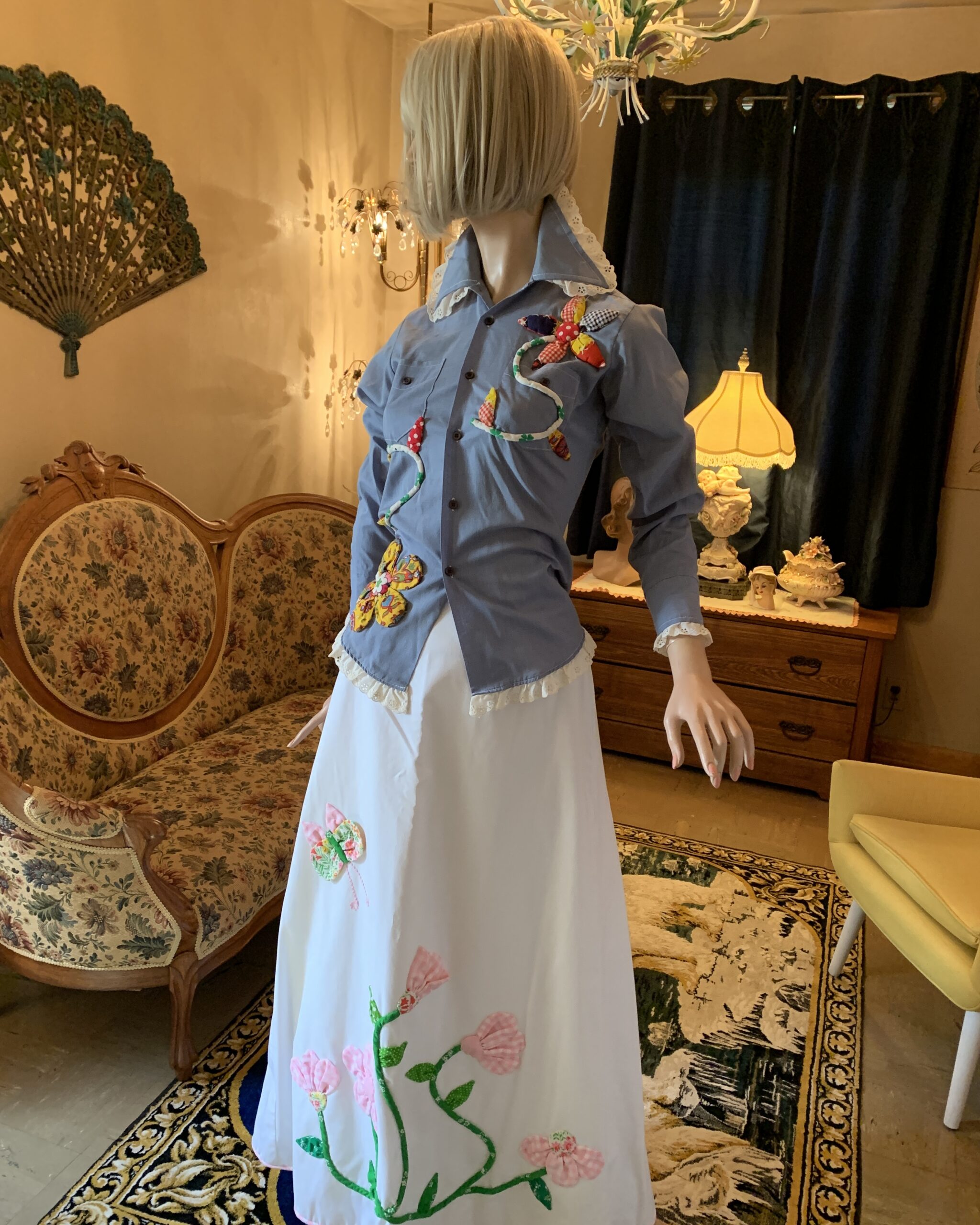 Angled view of a vintage Catherine Carr white maxi skirt featuring handcrafted floral appliqués, trailing green vines, and a butterfly accent.