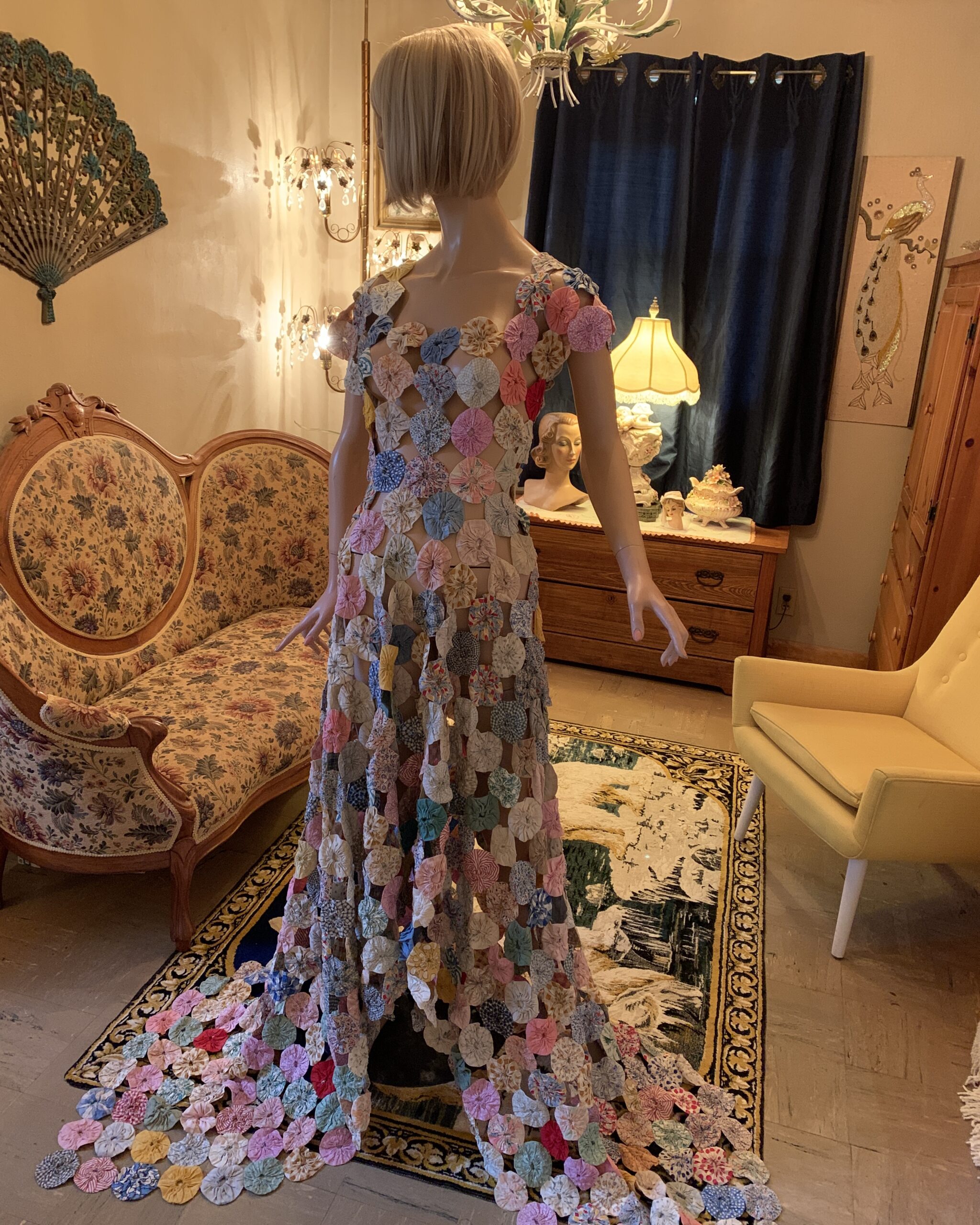 Full-length handmade yo-yo quilt dress displayed on a mannequin, featuring hundreds of fabric yo-yos arranged in an open lattice pattern with a dramatic trailing hem.