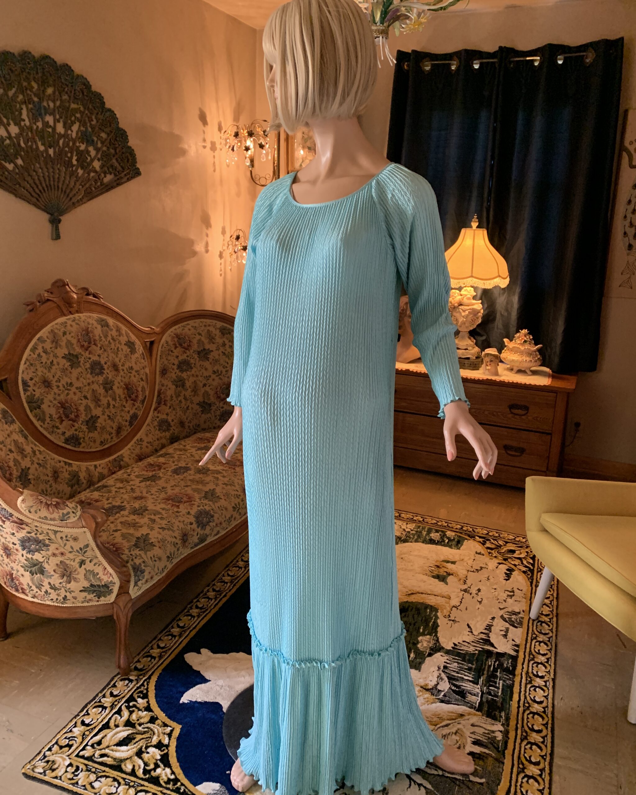 Vintage aqua pleated maxi gown displayed on a mannequin in a salon interior. The floor-length dress features a soft scoop neckline, three-quarter sleeves, an allover vertical pleated texture, and a flounced hem. Warm lamplight and antique furnishings create an elegant archival presentation.