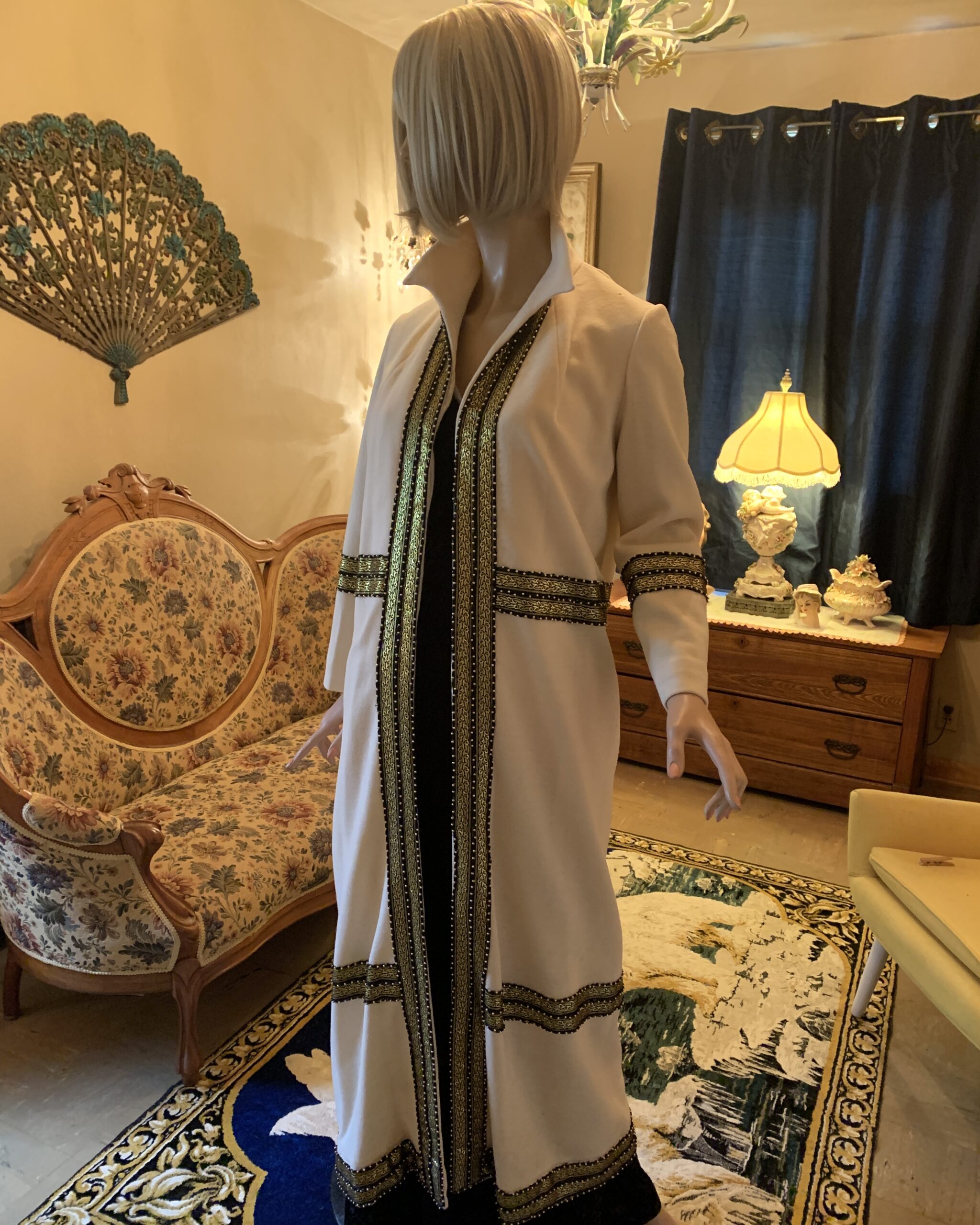 Vintage ivory opera coat with black and gold braided trim, standing collar, and full-length silhouette displayed in a salon setting.