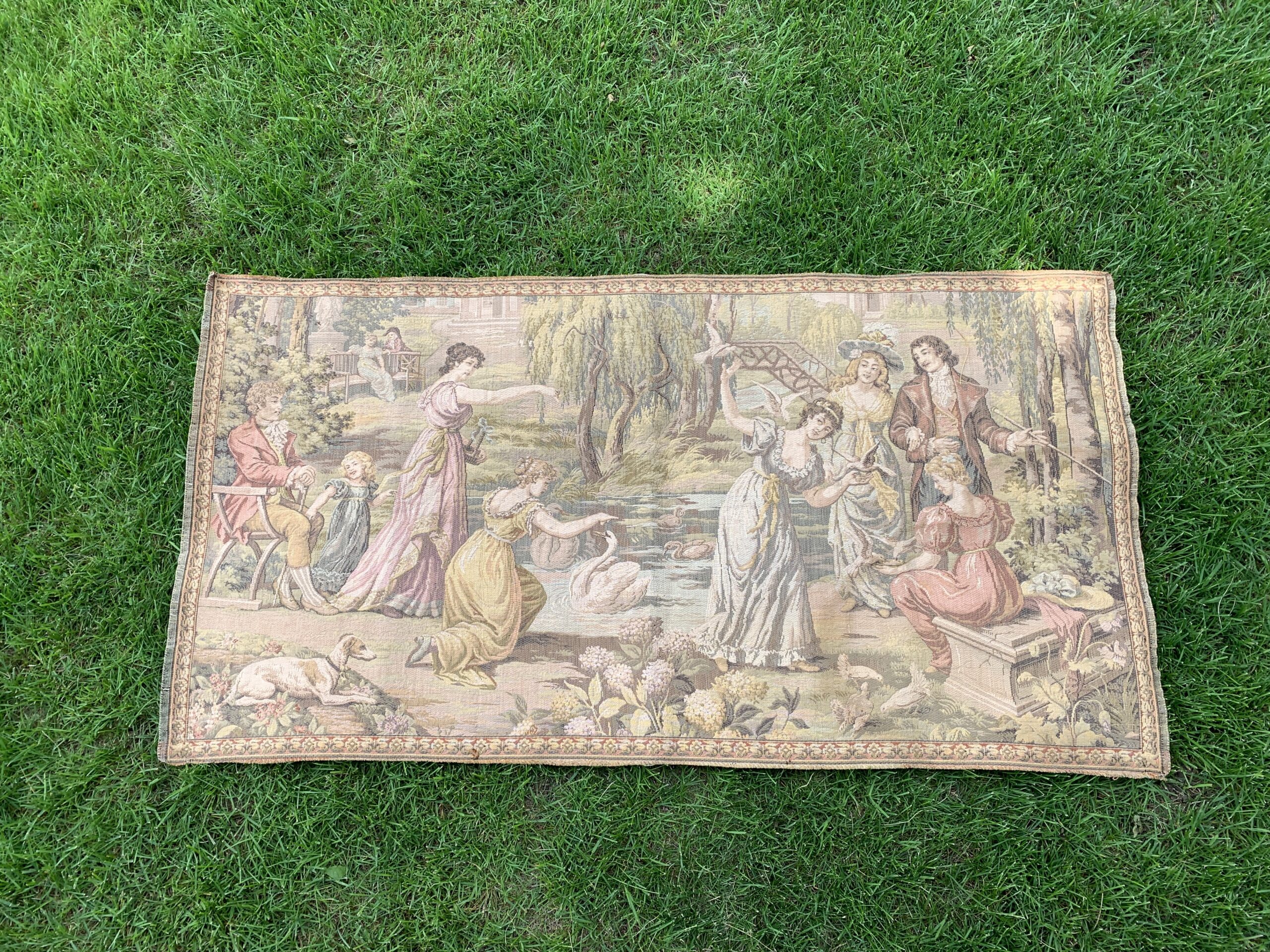 woven tapestry depicting elegantly dressed figures, swans, a pond, and a garden landscape photographed outdoors on green gras