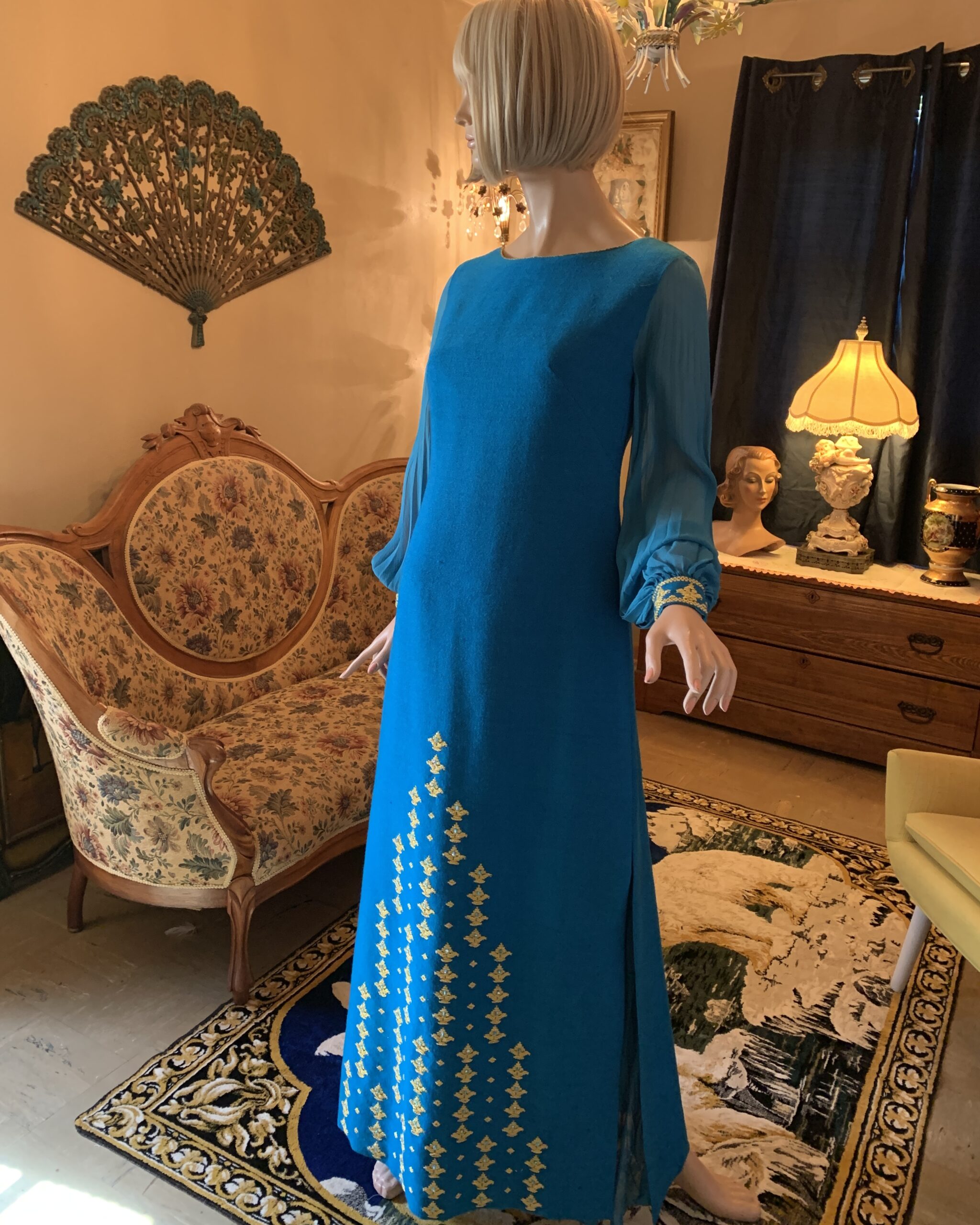 1970s Nikos and Takis Greek hostess gown in vibrant blue with gold metallic embroidery, sheer sleeves, and long column silhouette displayed on mannequin in salon interior.