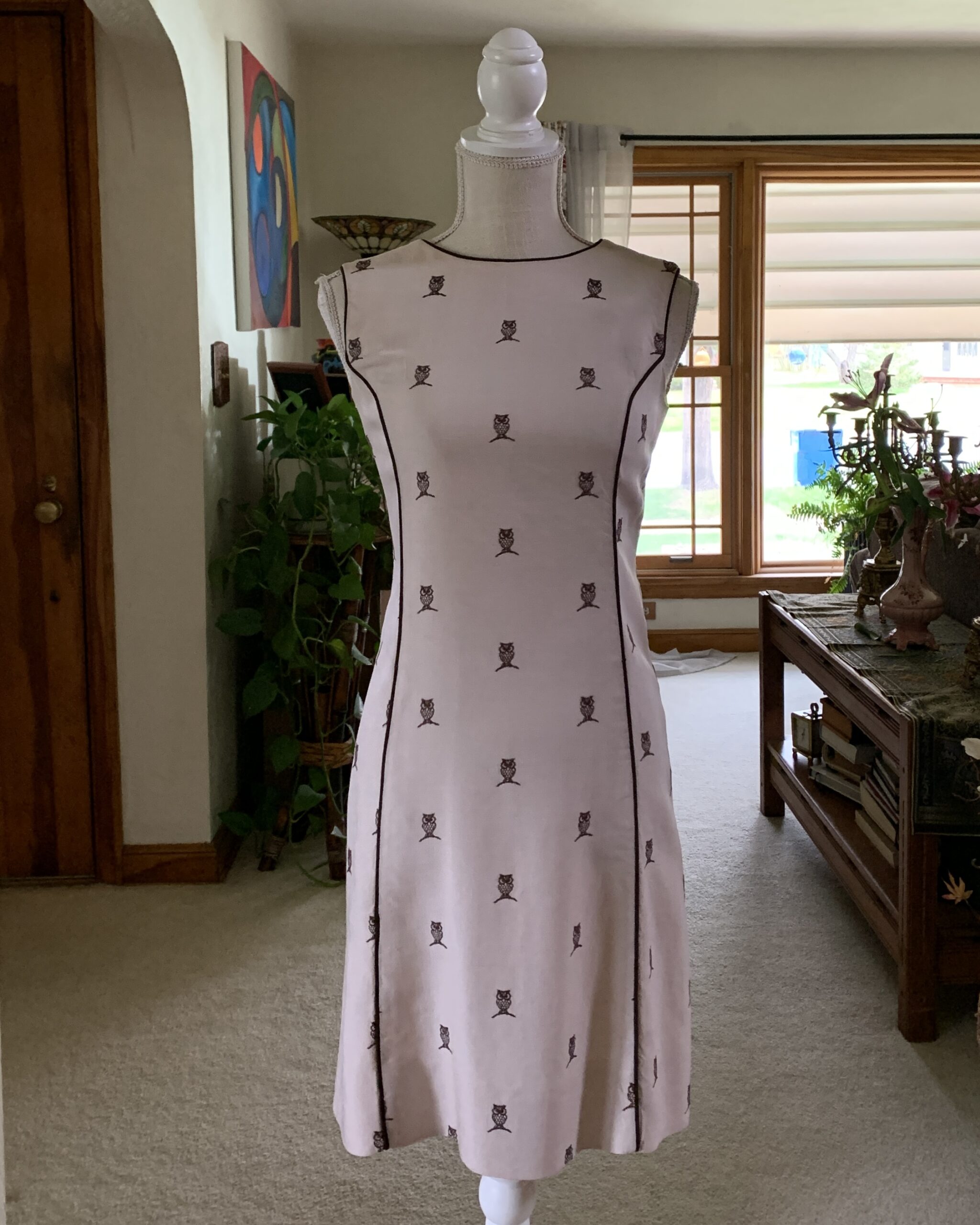 1960s Lord & Taylor linen owl print dress displayed on a mannequin beneath an interior archway, featuring a soft blush beige tone, contrast piping, sleeveless silhouette, and scattered miniature owl