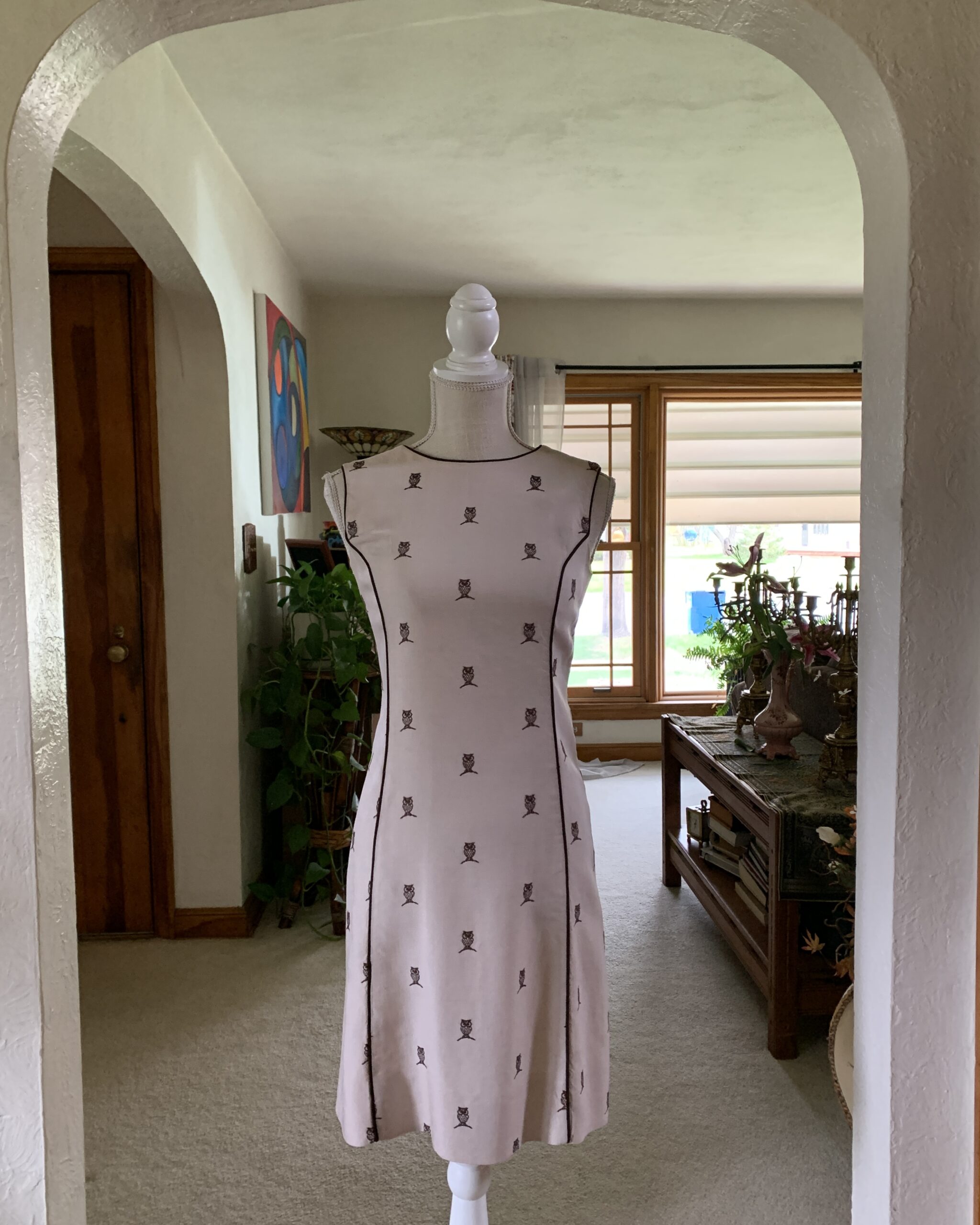 1960s Lord & Taylor linen owl print dress displayed on a mannequin beneath an interior archway, featuring a soft blush beige tone, contrast piping, sleeveless silhouette, and scattered miniature owl motifs.