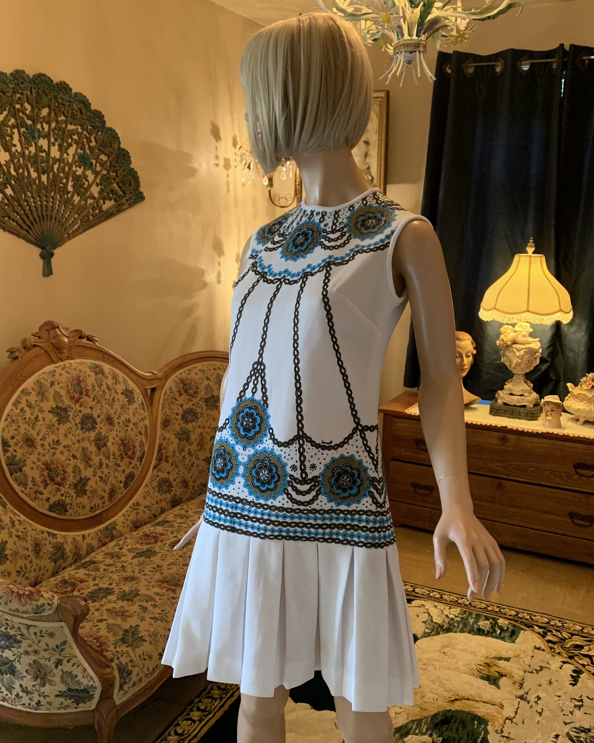 Side view of a 1960s Bernardo signed white dress featuring turquoise floral medallions, black decorative trim pattern, and a softly pleated drop-waist skirt.