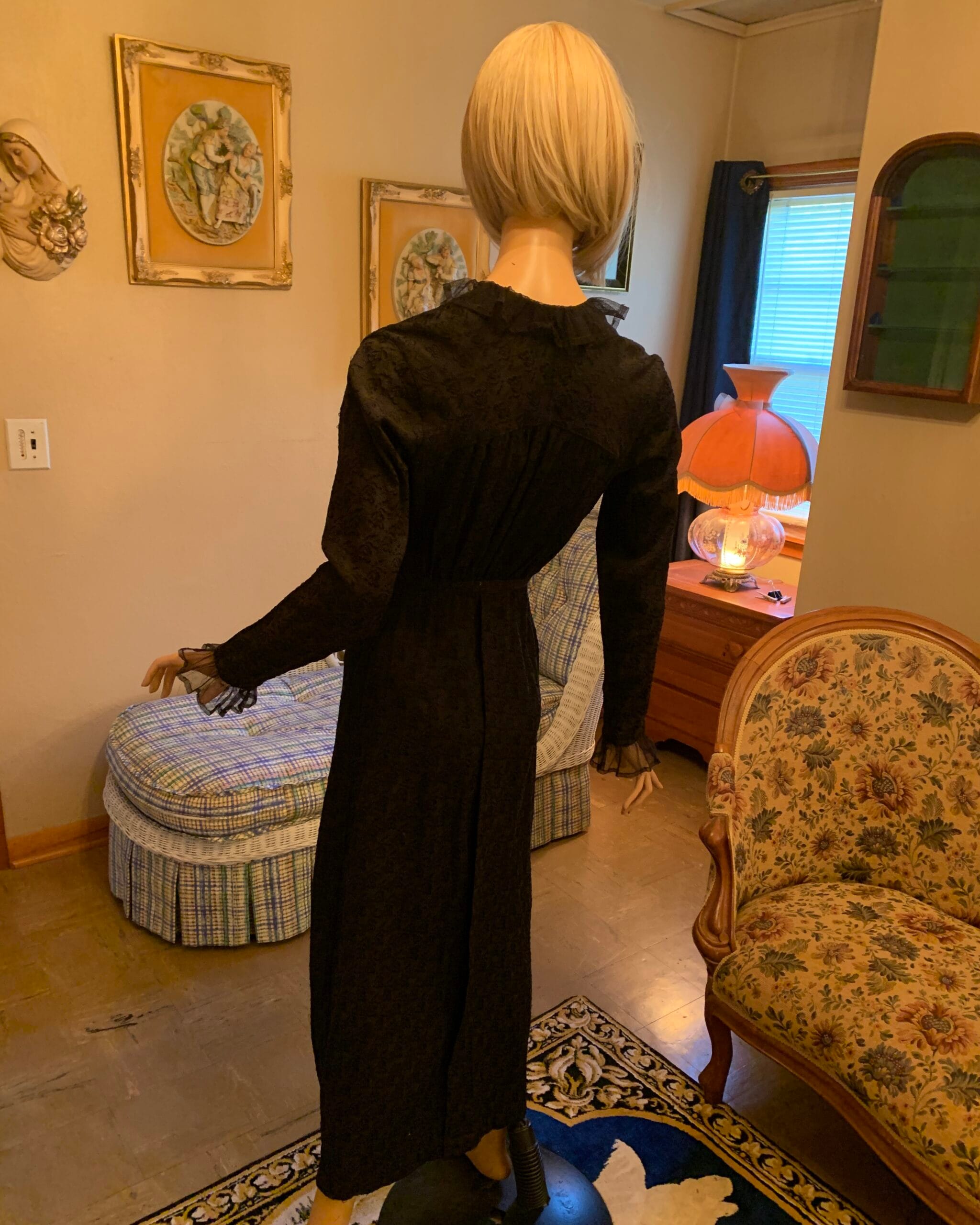 Back view of an antique black floral dress with gathered shoulders, fitted waist, and long skirt, displayed in a softly lit vintage interior.