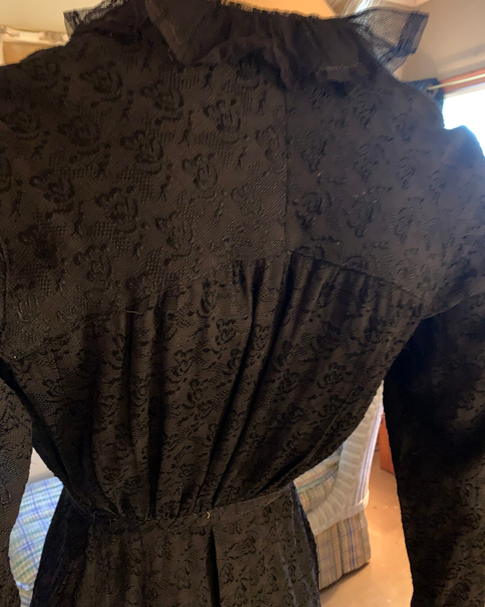 Rear shoulder detail of an antique black dress illuminated by warm lamplight, highlighting the floral woven fabric and gathered construction.