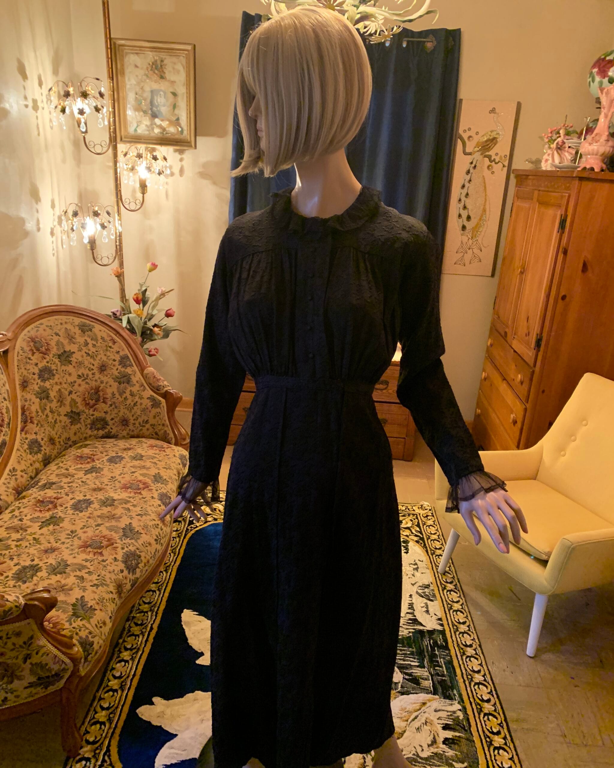 Antique black floral woven dress displayed in a vintage salon, featuring a ruffled neckline, covered front buttons, fitted waist, and long sleeves with sheer lace cuffs.