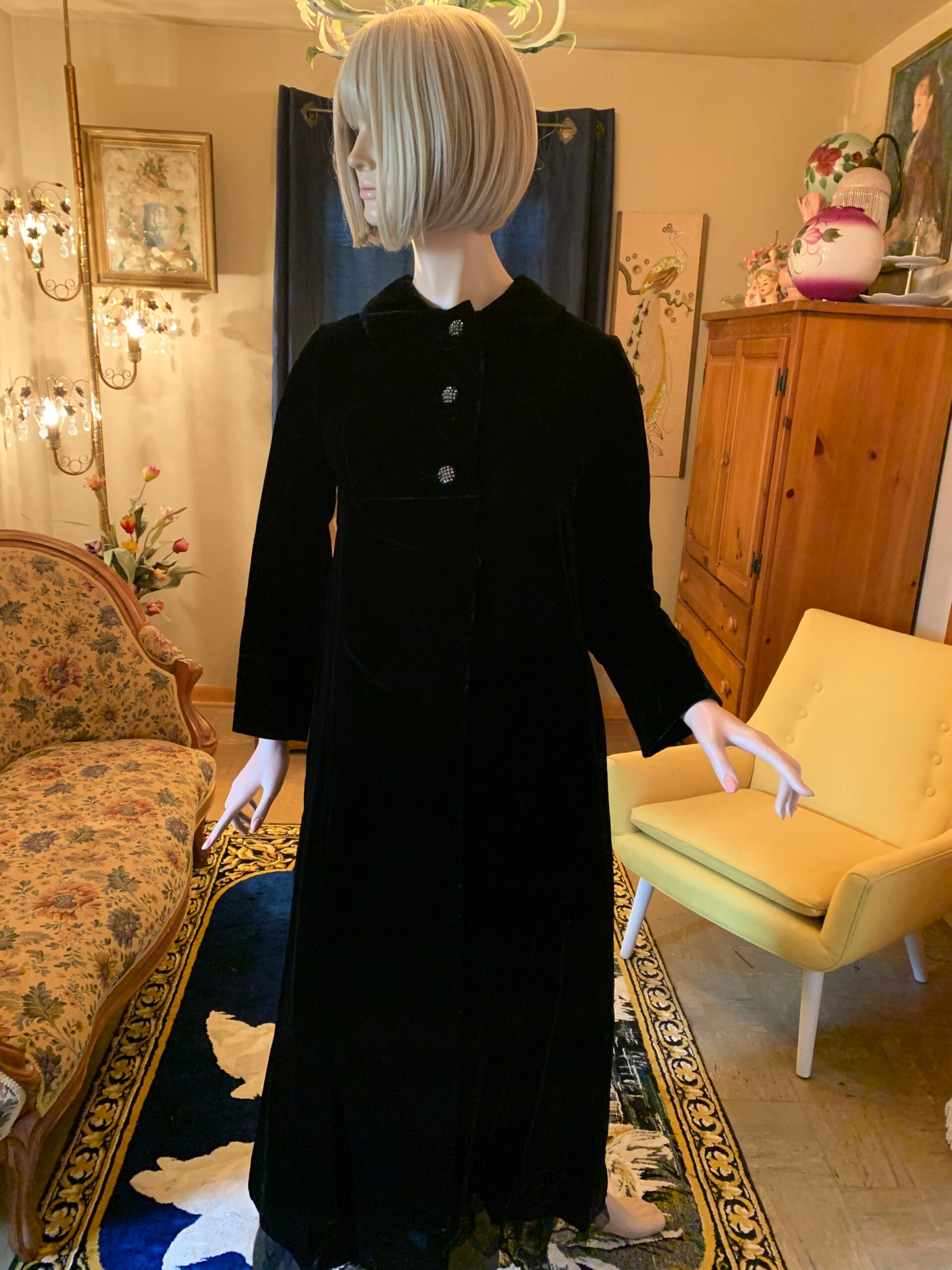 Black velvet opera coat with rhinestone buttons displayed in the salon room beside a floral settee and vintage furnishings.