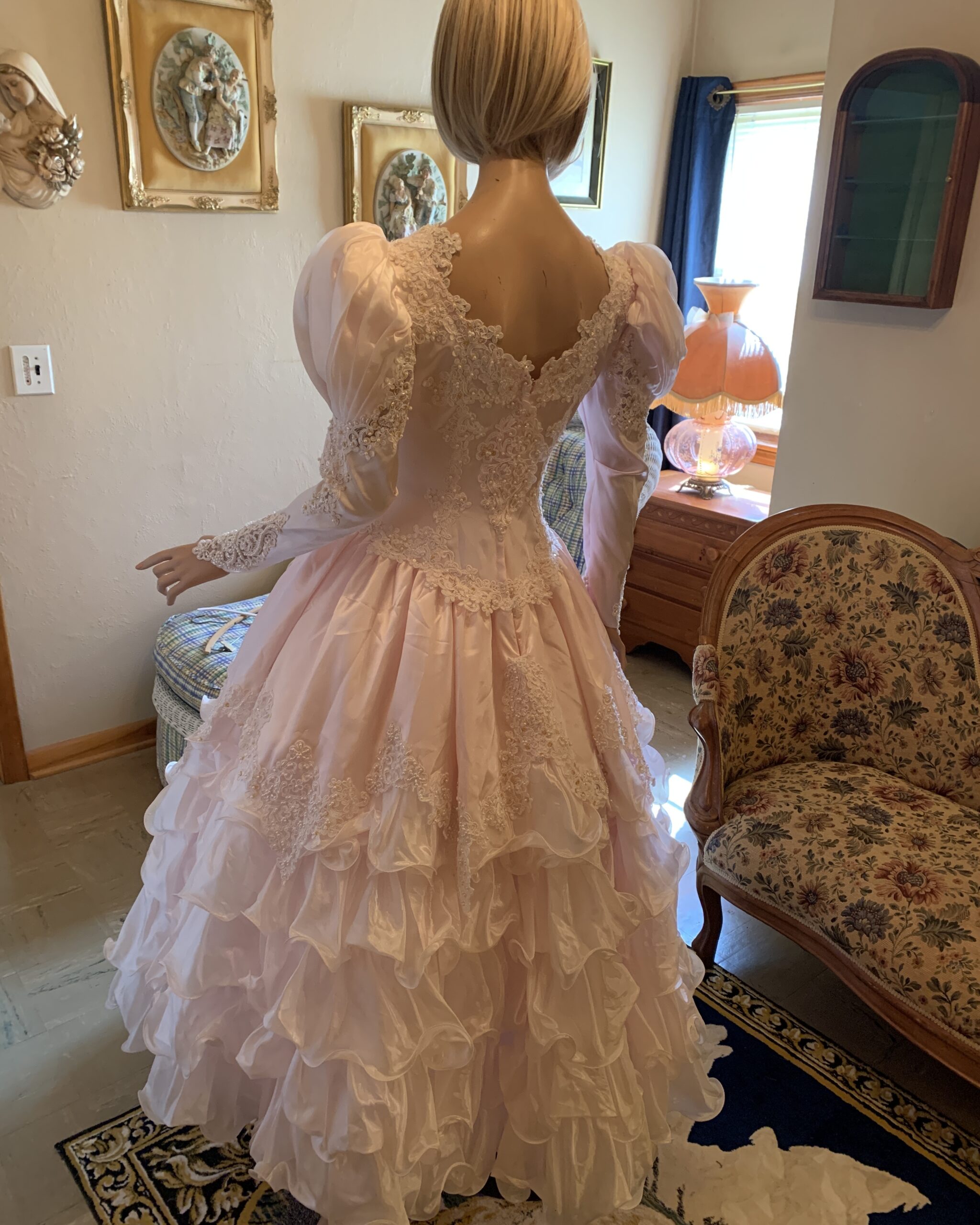 view of a vintage blush pink bridal gown featuring a lace embellished illusion back, dramatic puff sleeves, fitted waist, and cascading tiered ruffle skirt displayed on a mannequin.