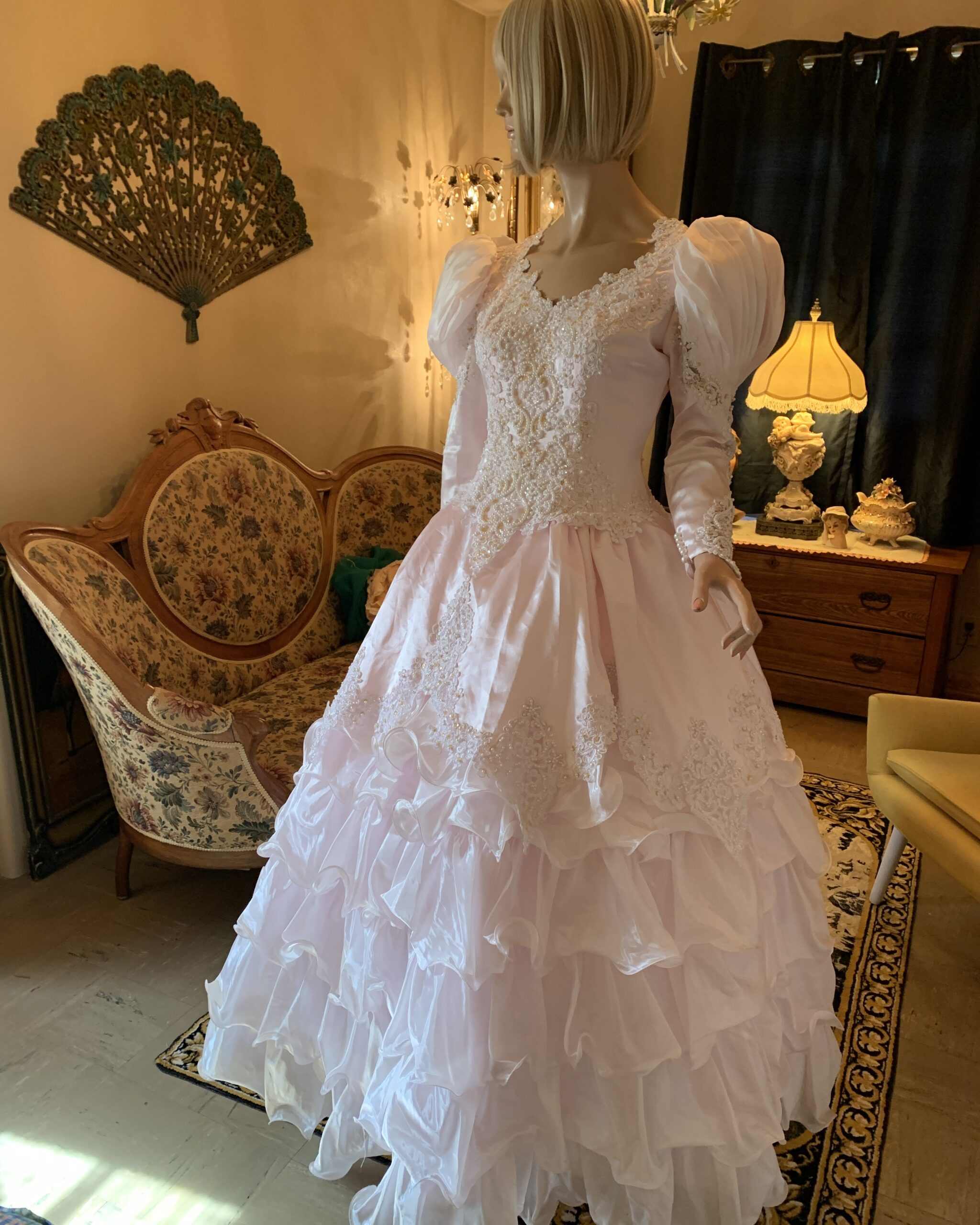 Vintage blush pink bridal gown with intricate lace and pearl embellished bodice, long puff sleeves, and dramatic tiered ruffle skirt displayed on a mannequin in a vintage-inspired salon interior.
