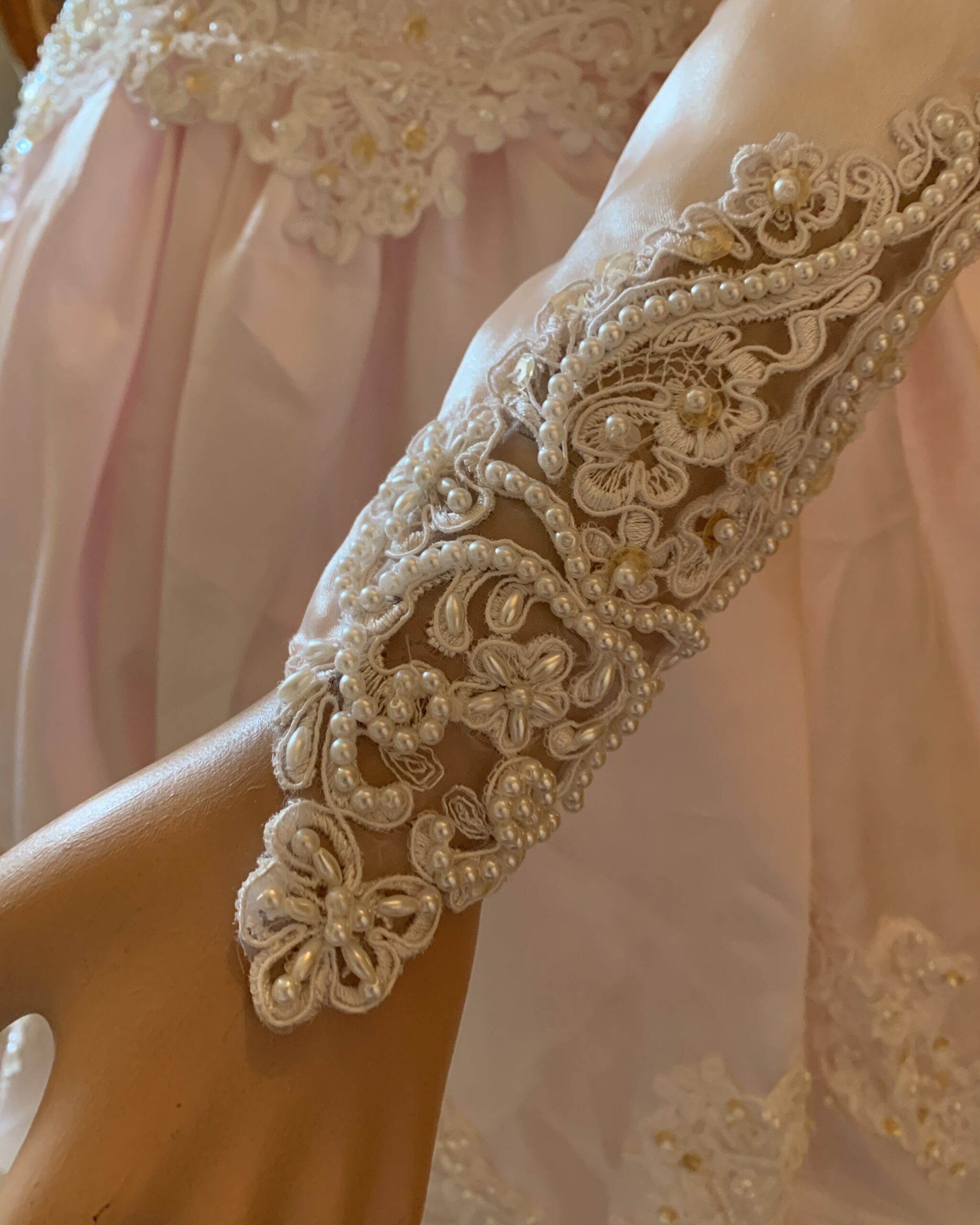 Close-up detail of a vintage blush pink bridal gown sleeve featuring intricate floral lace appliqué, faux pearl embellishments, and sheer illusion panels at the cuff.