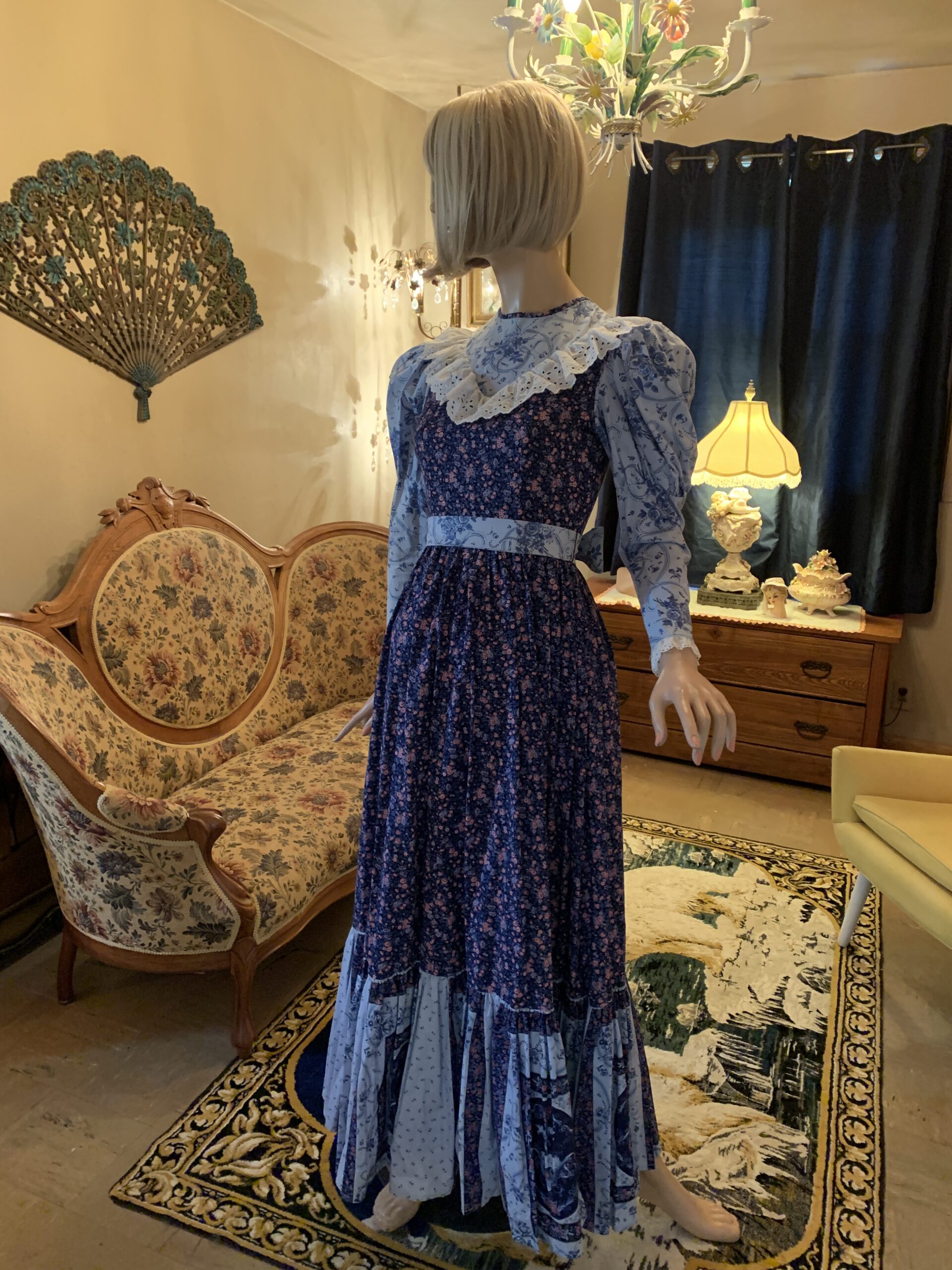 Handmade blue patchwork prairie dress from The Seamstress Collection displayed on a mannequin in a vintage salon setting. The cotton dress features puffed shoulders, a floral print bodice and skirt, eyelet-trimmed yoke, contrasting blue print sleeves, and a patchwork hem.