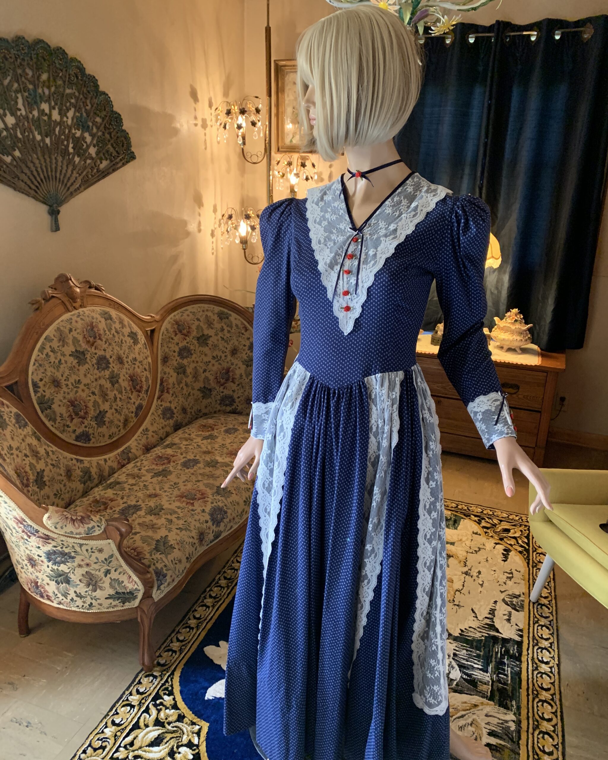 Blue Ribbon Prairie Dress from The Seamstress Collection featuring navy floral cotton, white lace bib, puff sleeves, ribbon rose accents, and full gathered skirt.