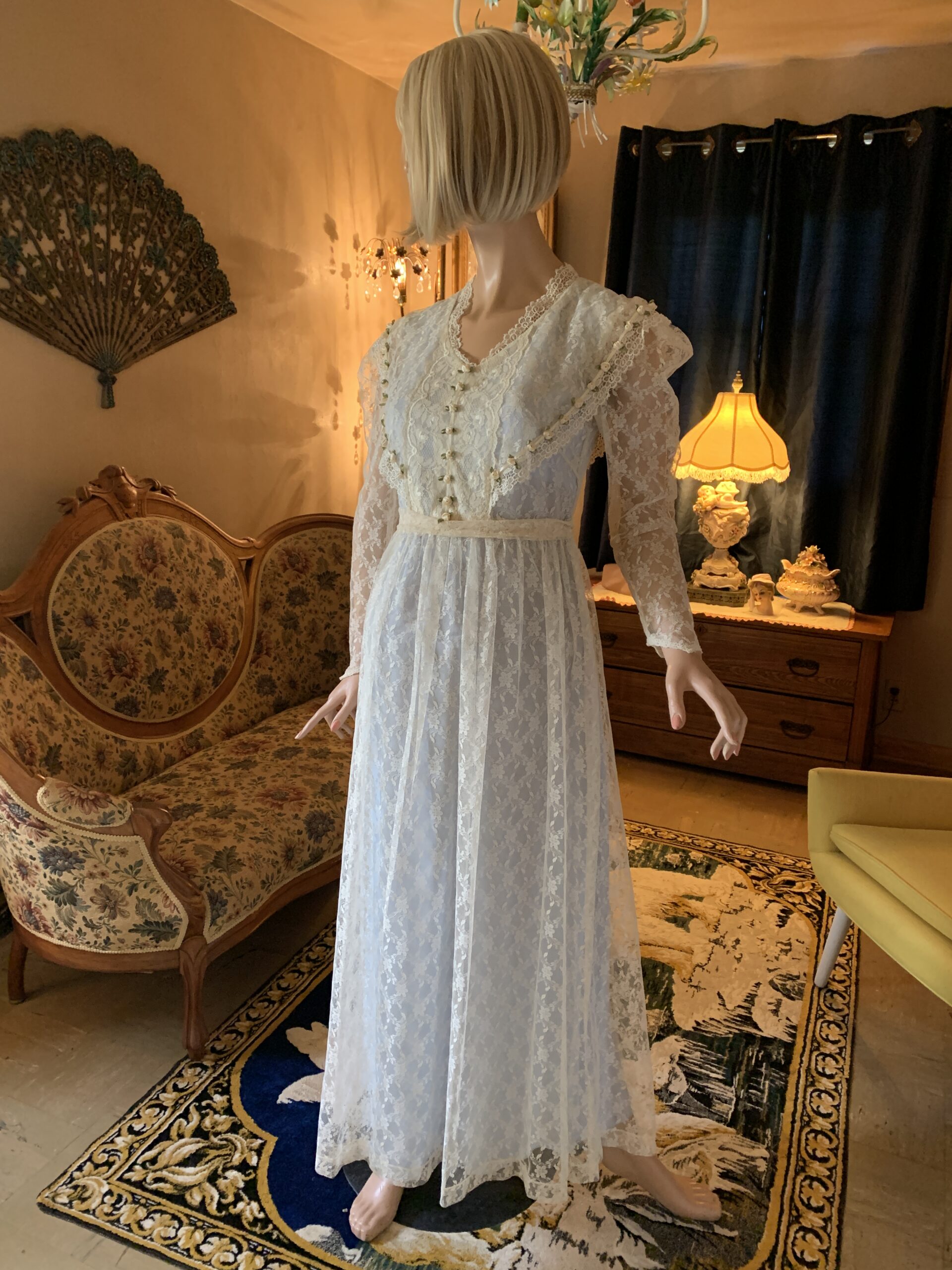 Handmade ivory lace gown from The Seamstress Collection featuring sheer sleeves, empire waist, ribbon rose trim, and floral lace overlay photographed in a vintage salon interior.