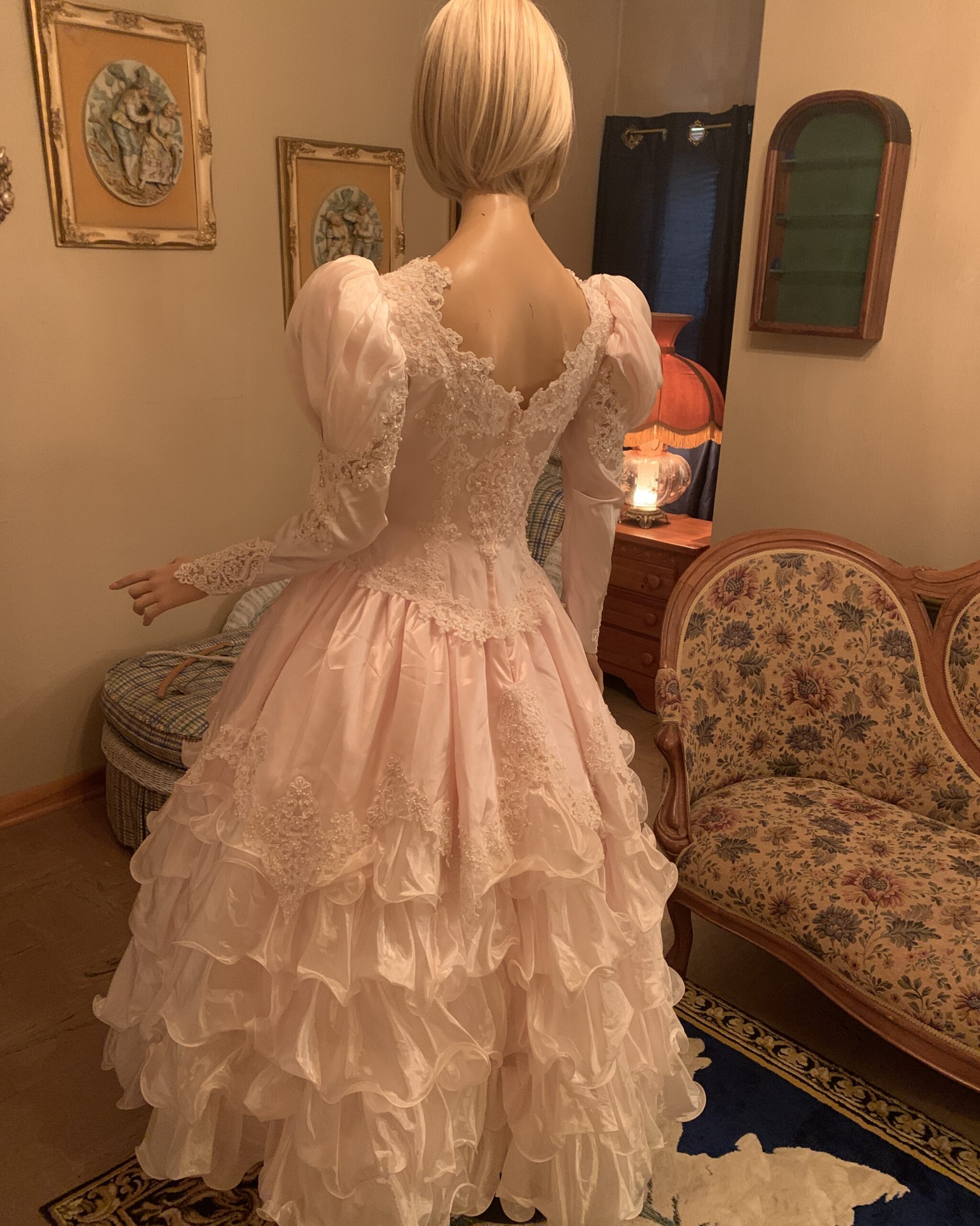 Back view of vintage blush pink bridal gown featuring illusion lace panel, pearl embellishment, puff sleeves, and full ruffled skirt.