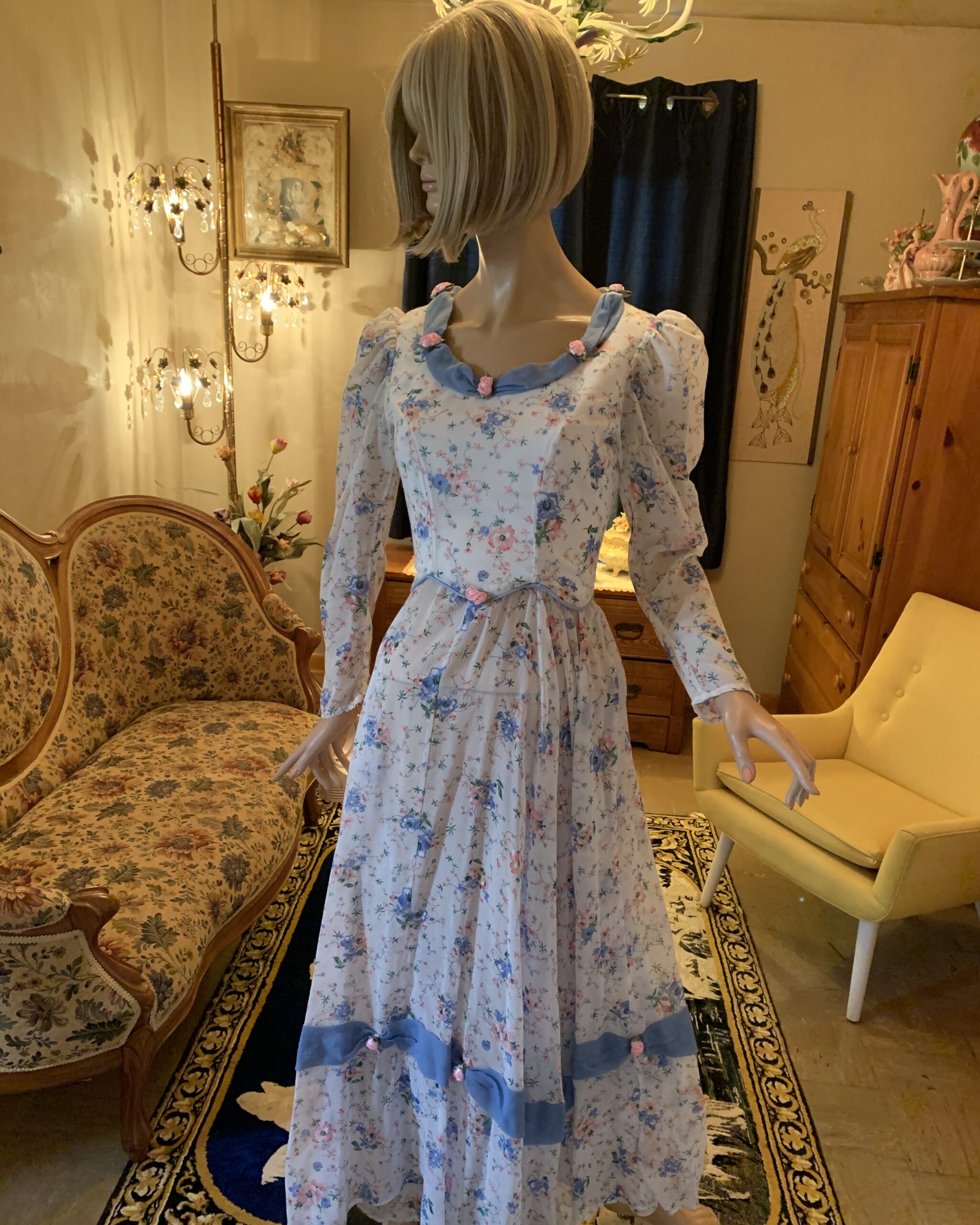Handmade western prairie dress in floral voile fabric with blue trim, ribbon rose accents, peplum waist, and long sleeves, photographed in the salon setting.