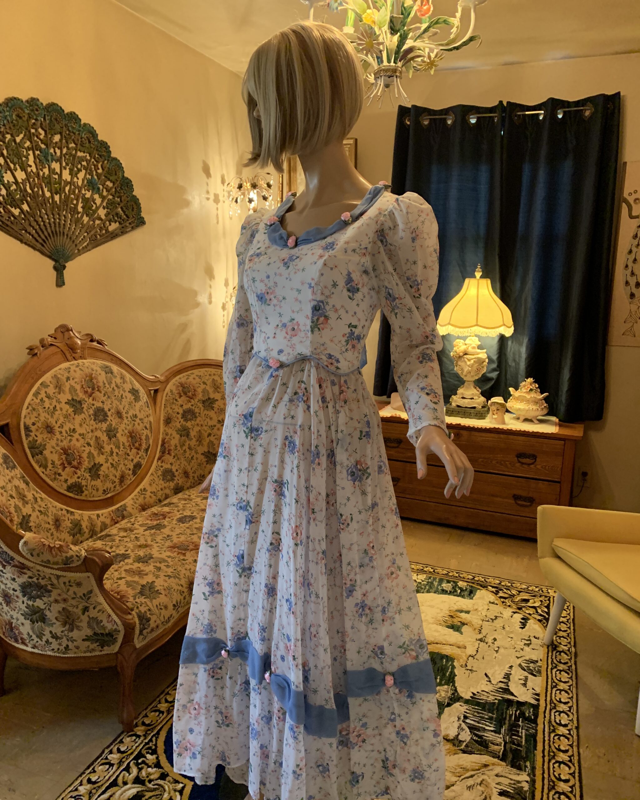 Three-quarter view of handmade floral prairie dress featuring blue trim, gathered skirt, peplum waist, and delicate ribbon rose embellishments.