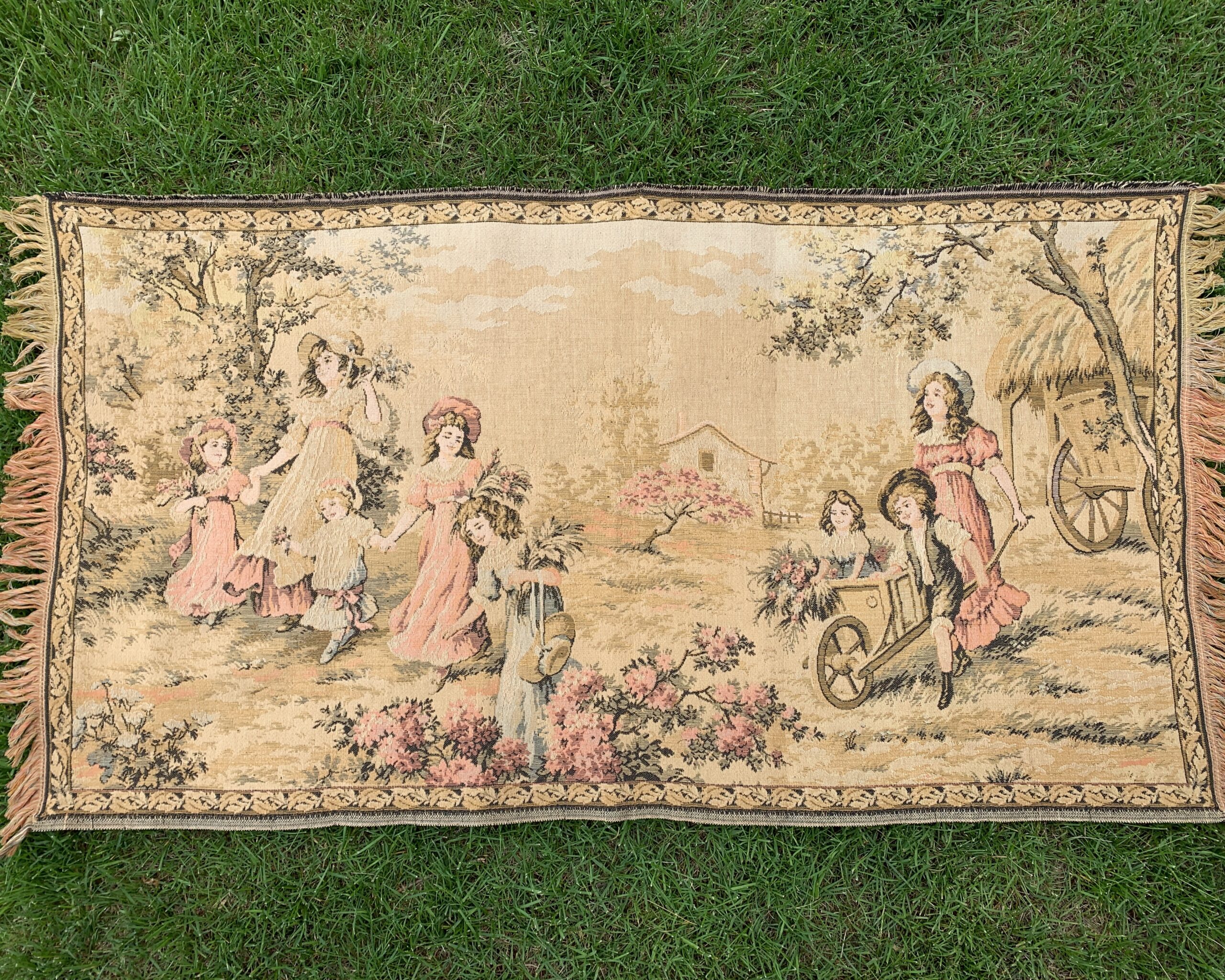 Vintage European woven wall hanging depicting a pastoral spring garden scene with women, children, flowering trees, and a floral border with multicolored fringe.