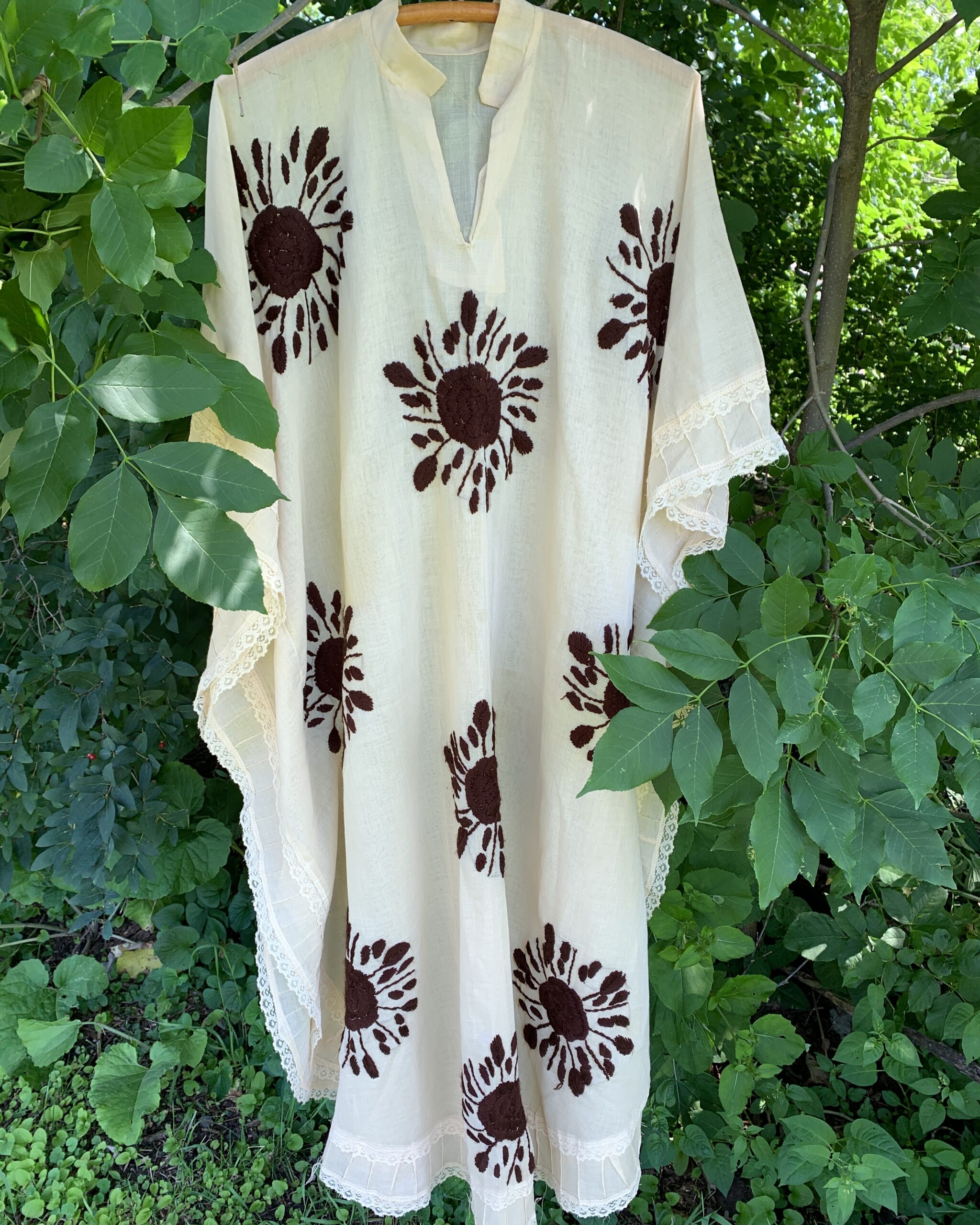 Vintage ivory caftan with hand-embroidered brown floral motifs, split neckline, lace-trimmed sleeves, pintucked hem, photographed outdoors in a garden.