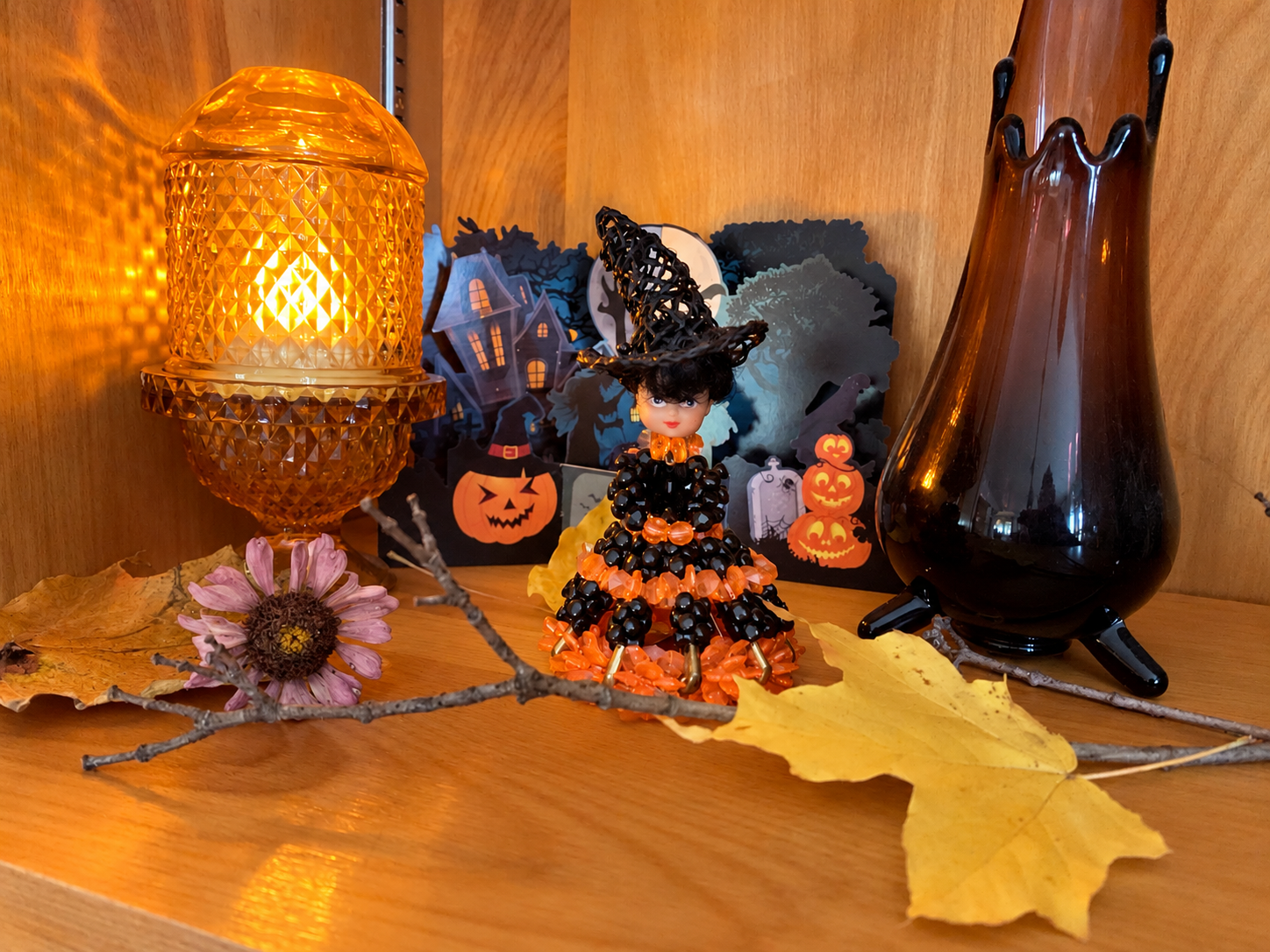 Vintage 1970s beaded witch doll displayed in the Halloween Cabinet beside glowing amber glass and black candleholders.