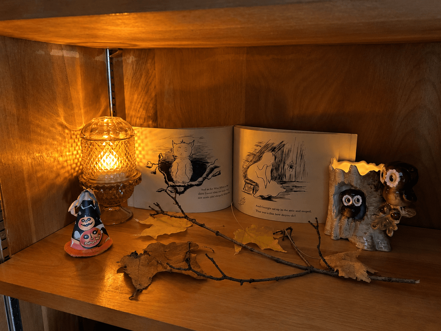 Open vintage Georgie the Ghost book displayed on a warm wooden Halloween shelf with amber lamp, autumn leaves, and owl figurines.