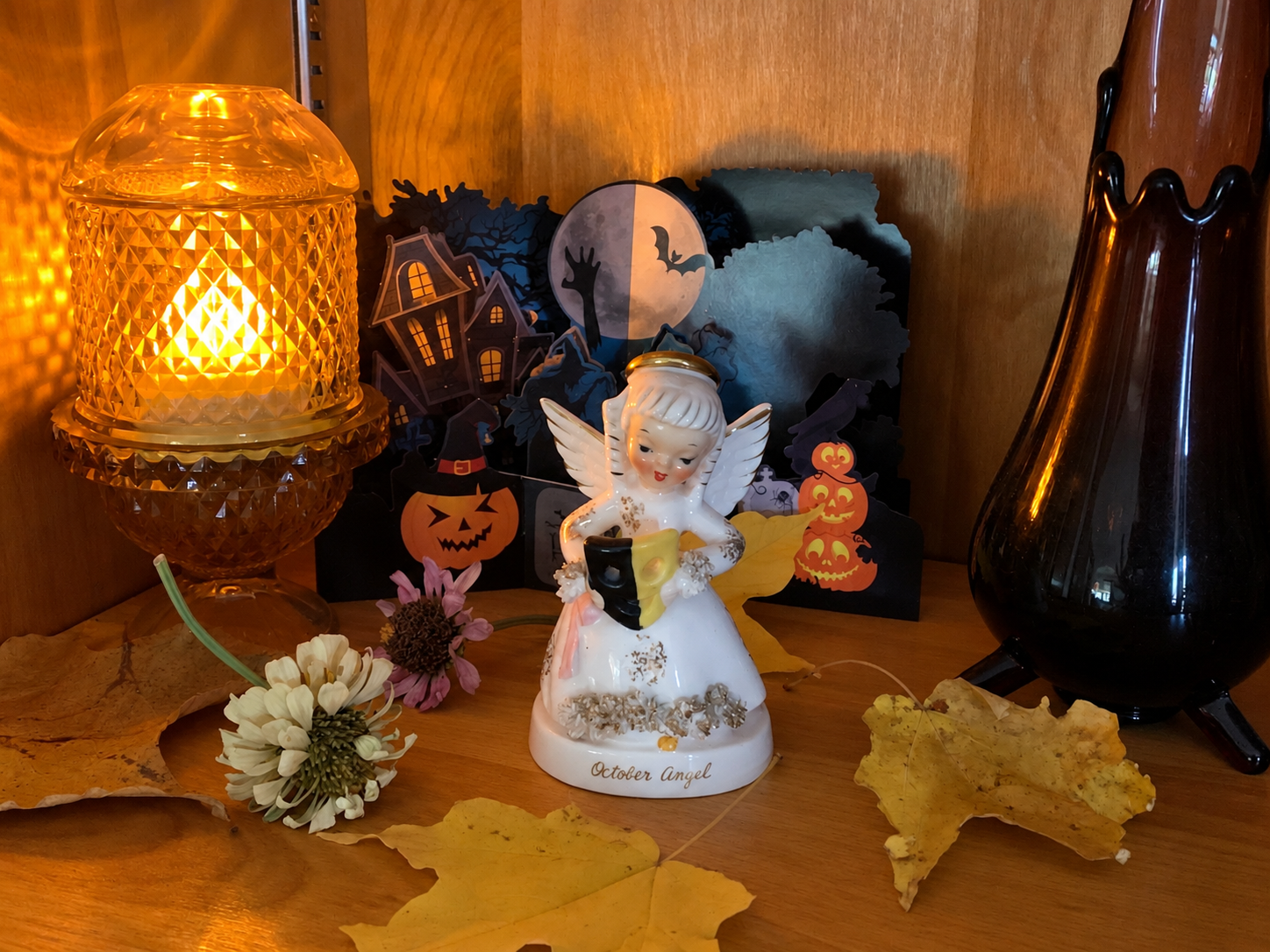 Vintage Napco October Angel figurine displayed in a warm wood cabinet beside an amber glass lamp, autumn leaves, and Halloween paper decorations.