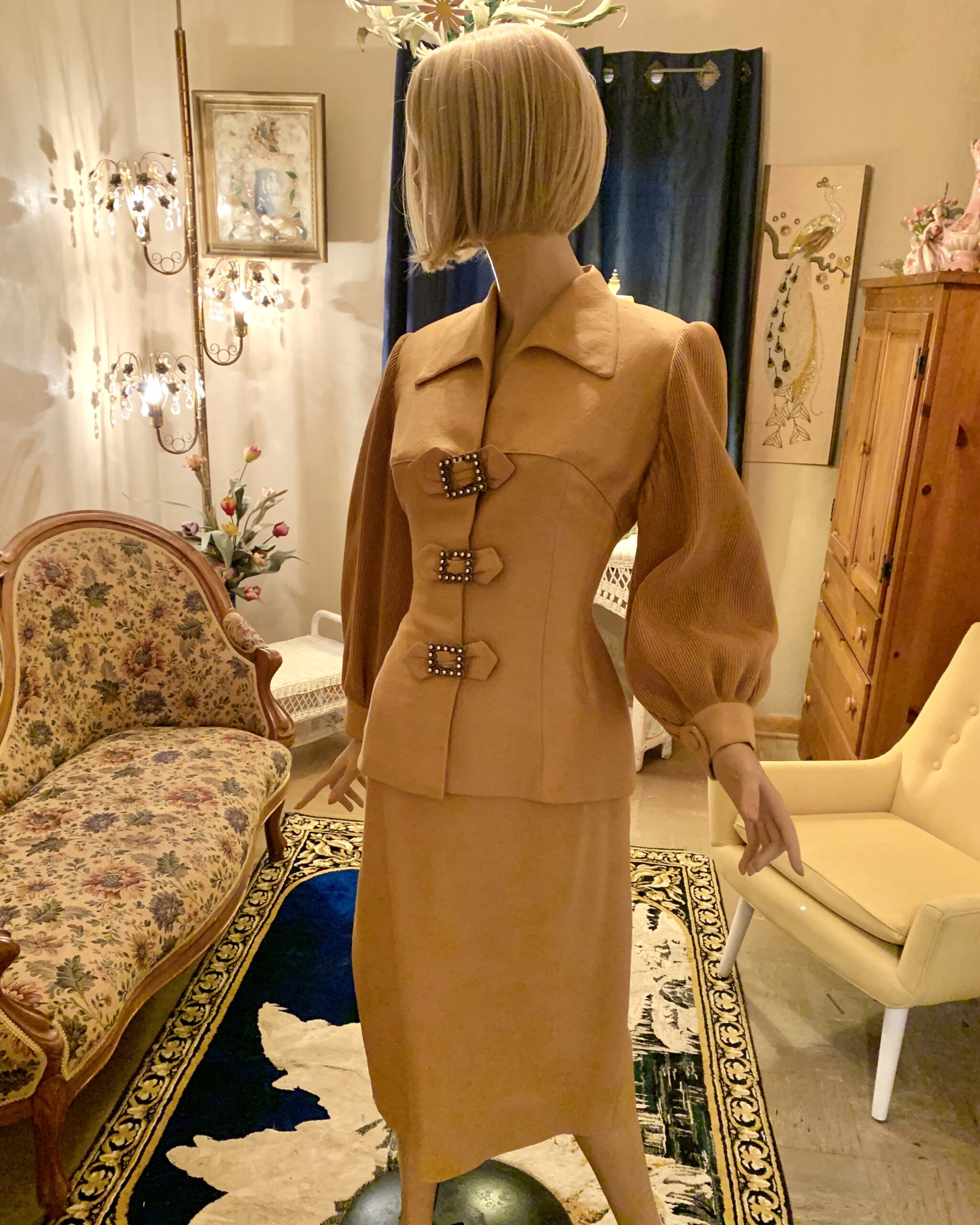 Vintage golden camel Lilli Ann skirt suit on a mannequin, with a fitted jacket, full pleated sleeves, and three rhinestone buckle accents.