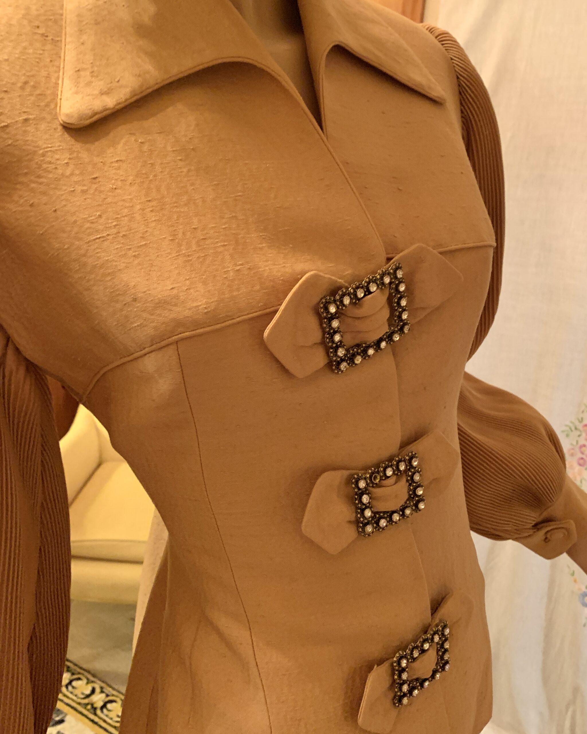 Close-up of the Lilli Ann jacket showing textured camel fabric, pleated sleeves, and three rectangular rhinestone buckle accents.