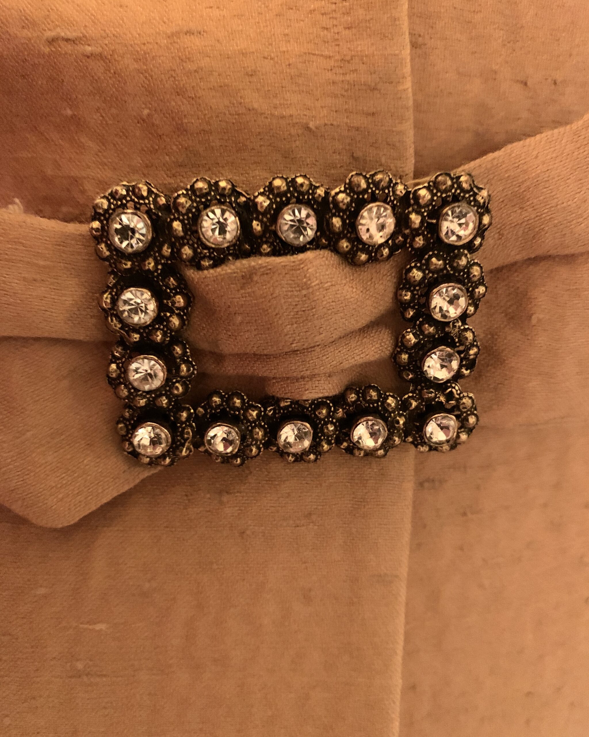 Close-up of a rectangular buckle with clear rhinestones and floral metalwork on a camel fabric tab.
