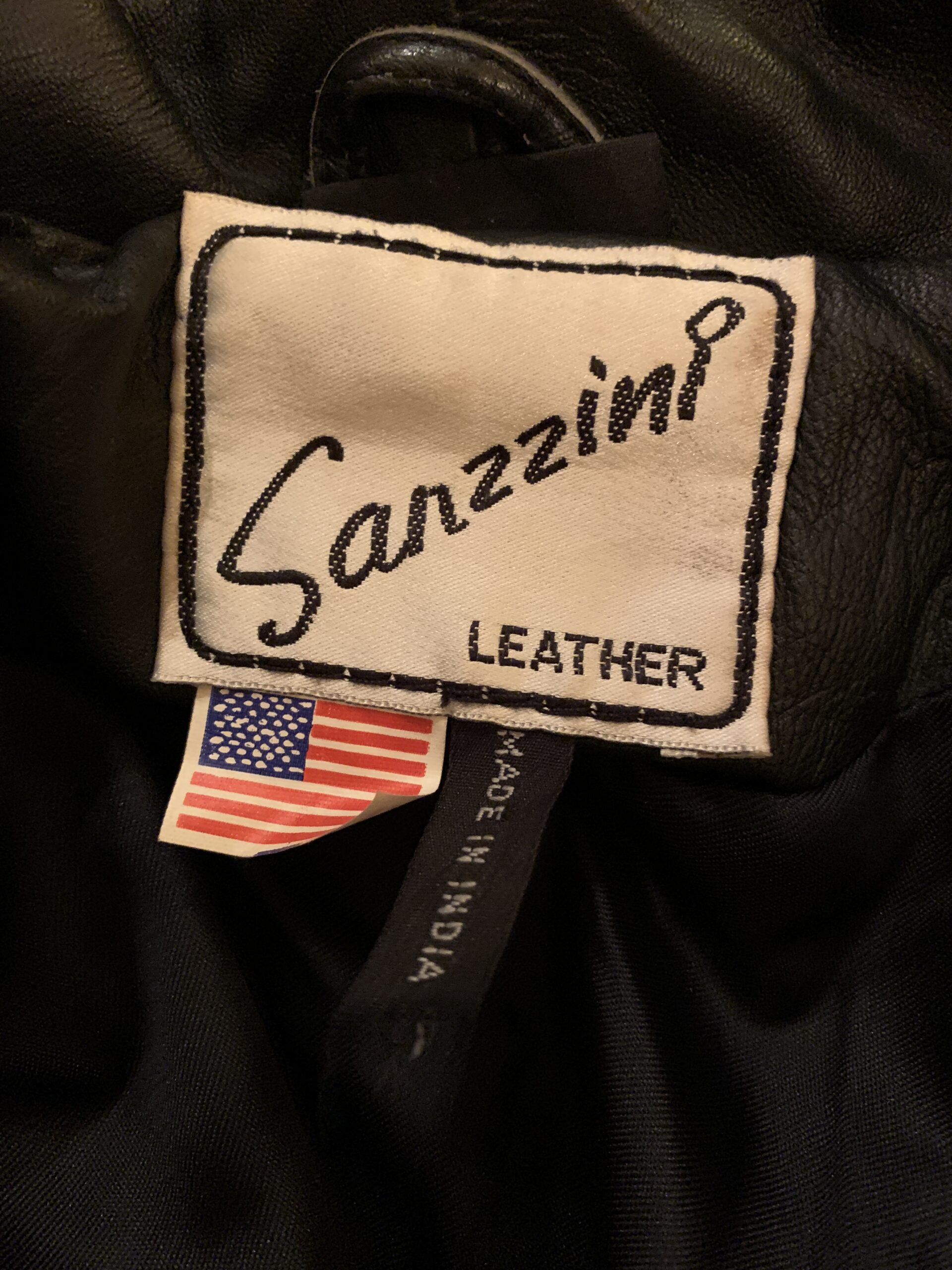 Close-up of the Sanzzini Leather designer label inside a vintage black leather cape coat, with American flag and Made in India tags.